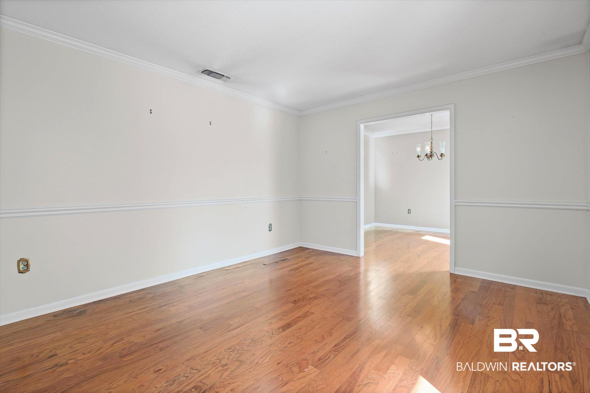 Vacant room with hardwood floors and chair rail molding, opening into a room with a chandelier.