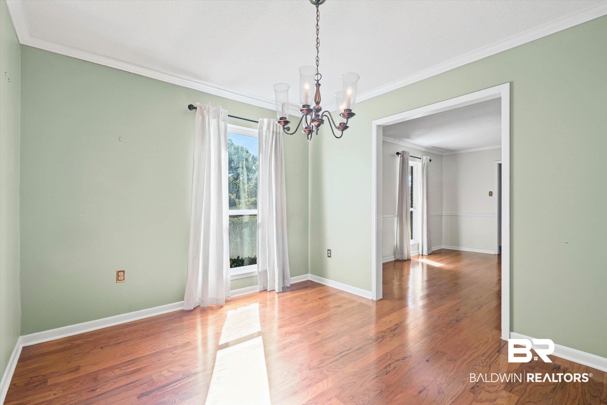 Empty dining room with pale green walls, hardwood floors, chandelier, and a wide doorway to another room.