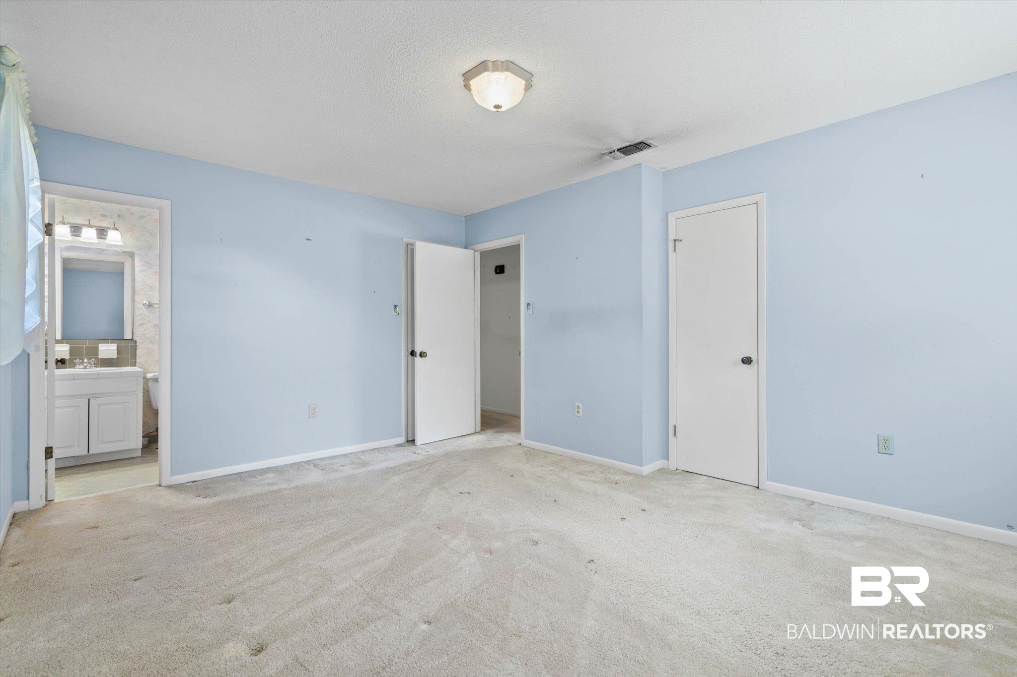 Empty bedroom with pale blue walls, worn carpet, and an open doorway to the bathroom.