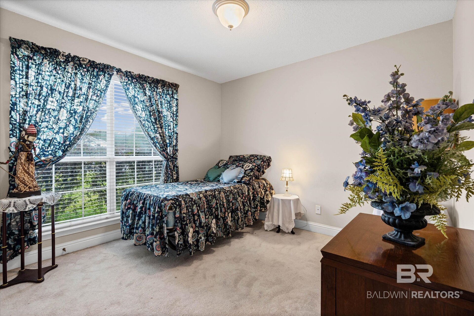 Bedroom with floral bedding and curtains, a large window, and a wooden dresser topped with blue flowers.