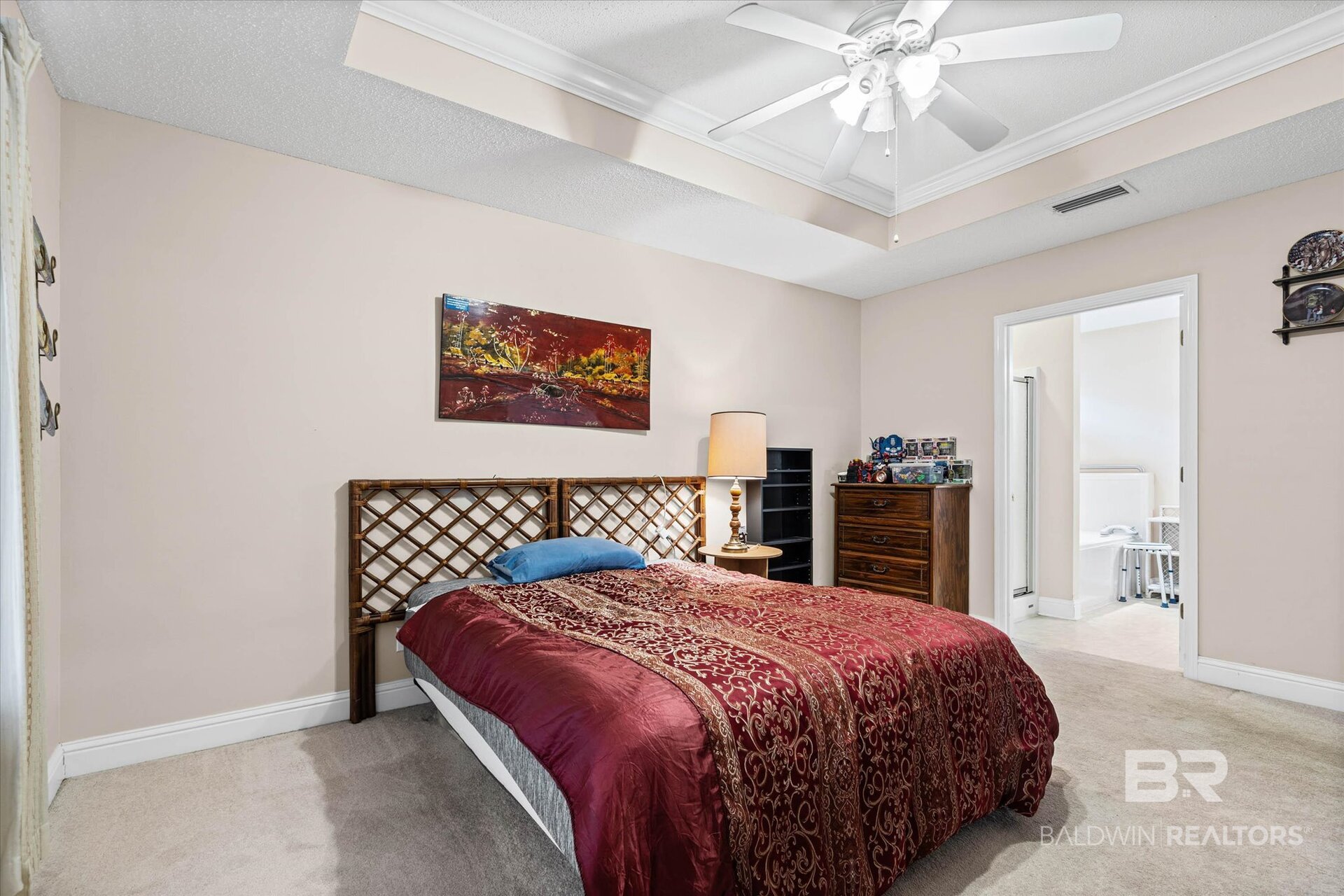 Furnished bedroom with a red patterned bedspread, lattice headboard, tray ceiling, and open bathroom doorway.