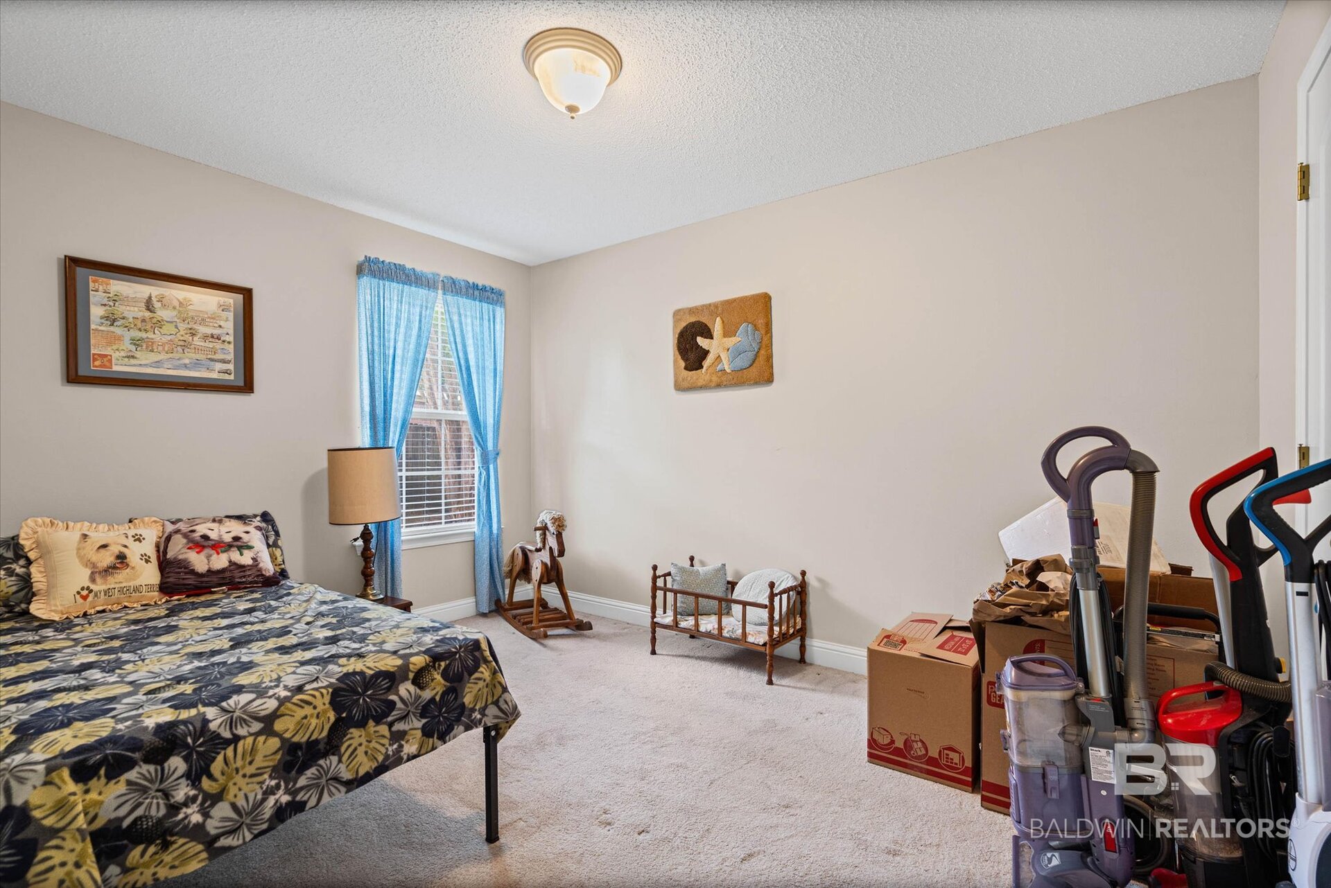 Bedroom with a patterned bed, blue curtains, and moving boxes and vacuums along one side.