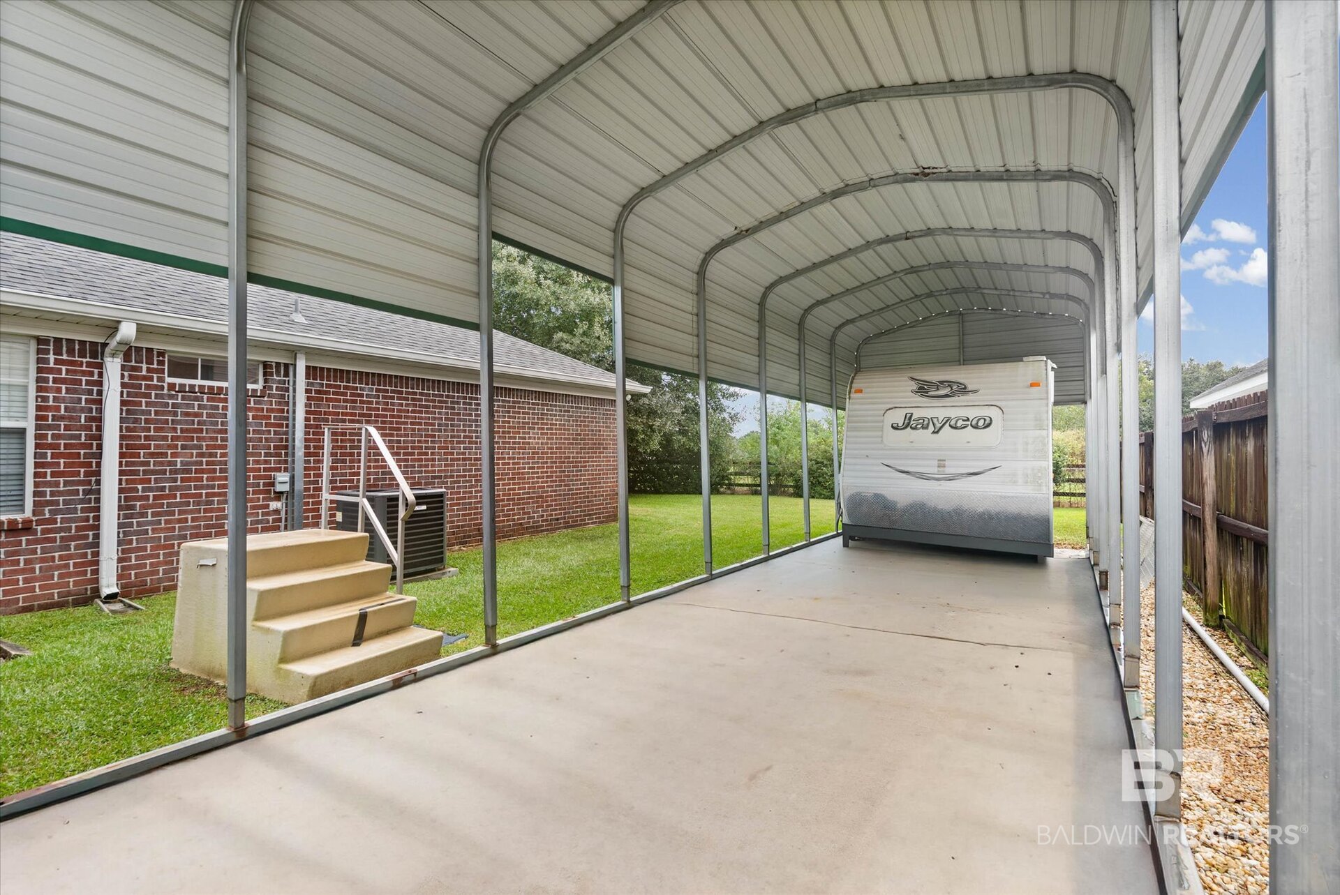 Covered RV carport beside a brick house, with a Jayco trailer parked on the concrete pad.