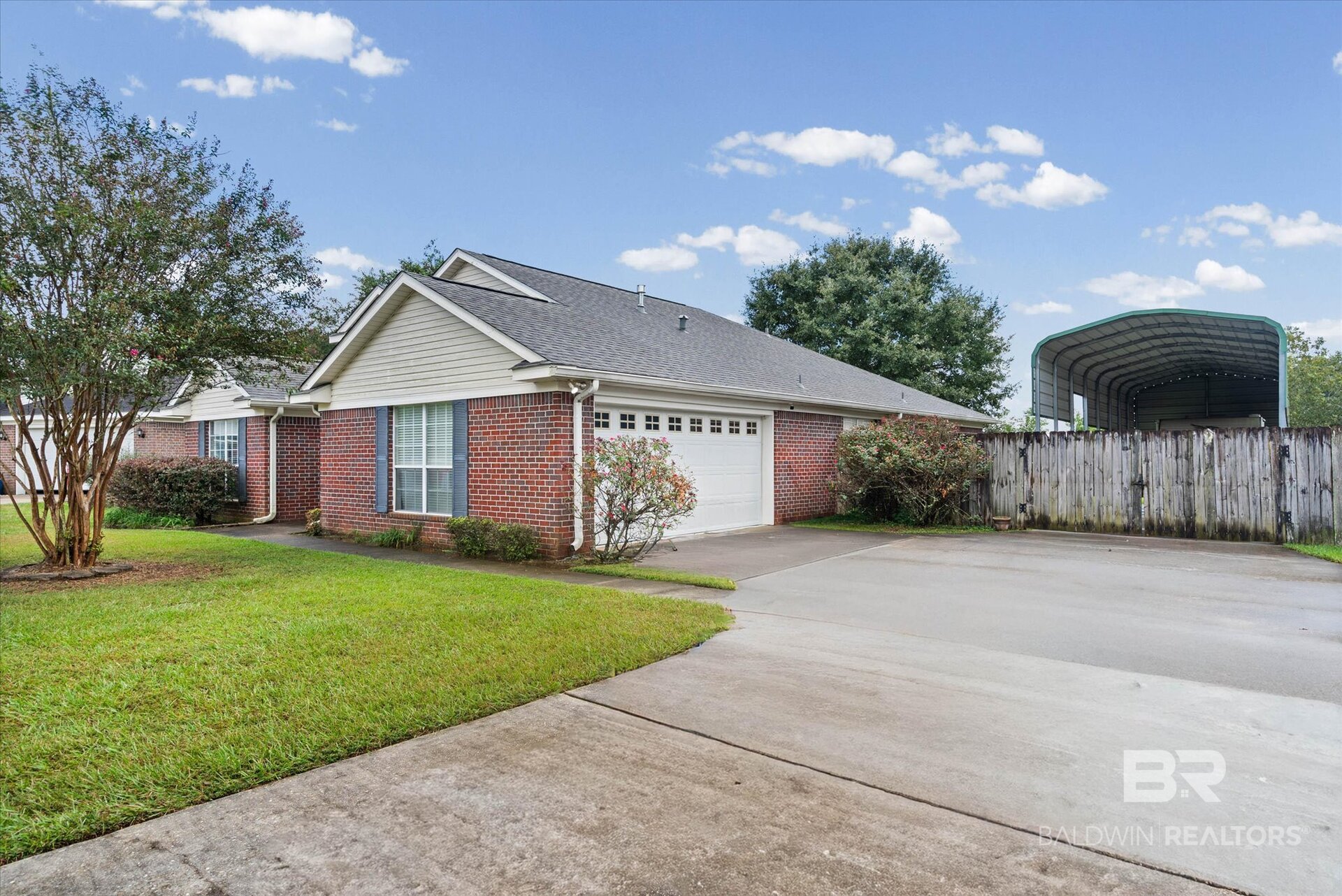 Single-story brick home with an attached double garage, broad driveway, and metal-roofed shelter behind a fence.