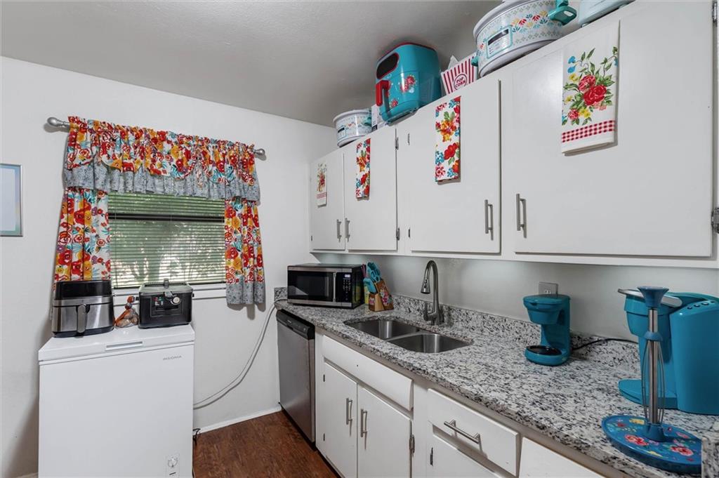 Compact kitchen with white cabinets, granite counters, a double sink, and a chest freezer beneath a curtained window.