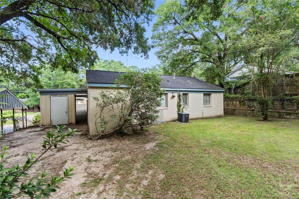 Rear of a single-story brick house beneath mature trees, with a covered side entrance and spacious yard.