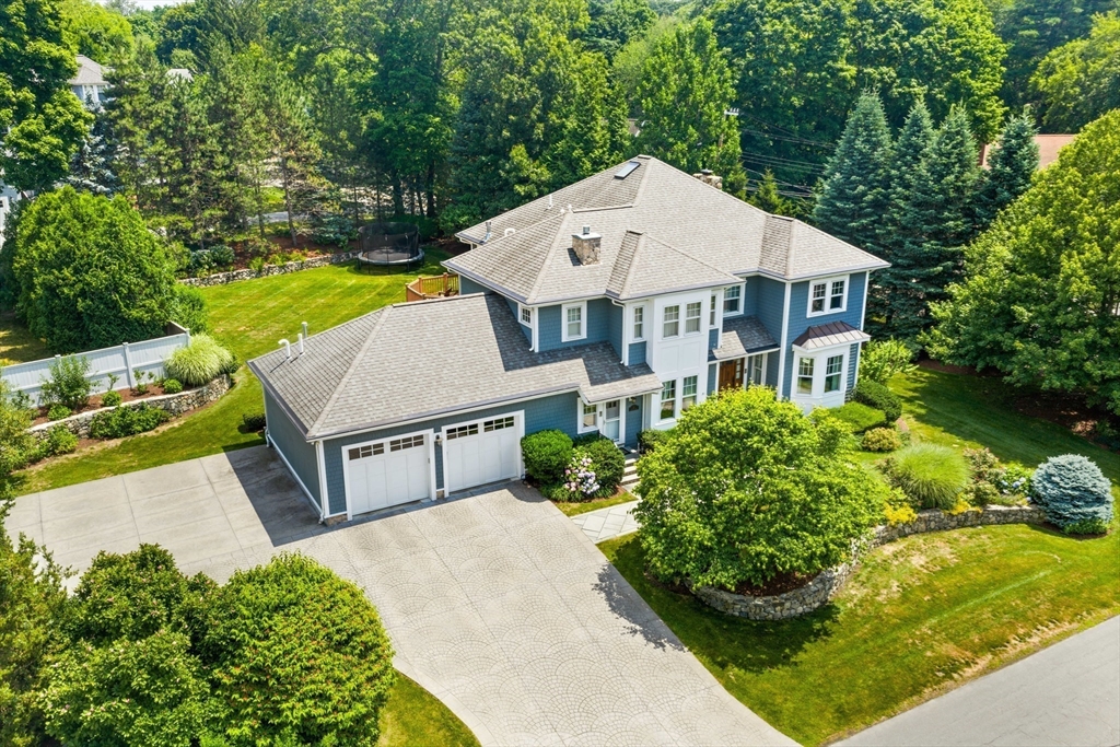 Aerial view of a blue-gray house with an attached three-car garage, landscaped lawn, and wooded backyard.