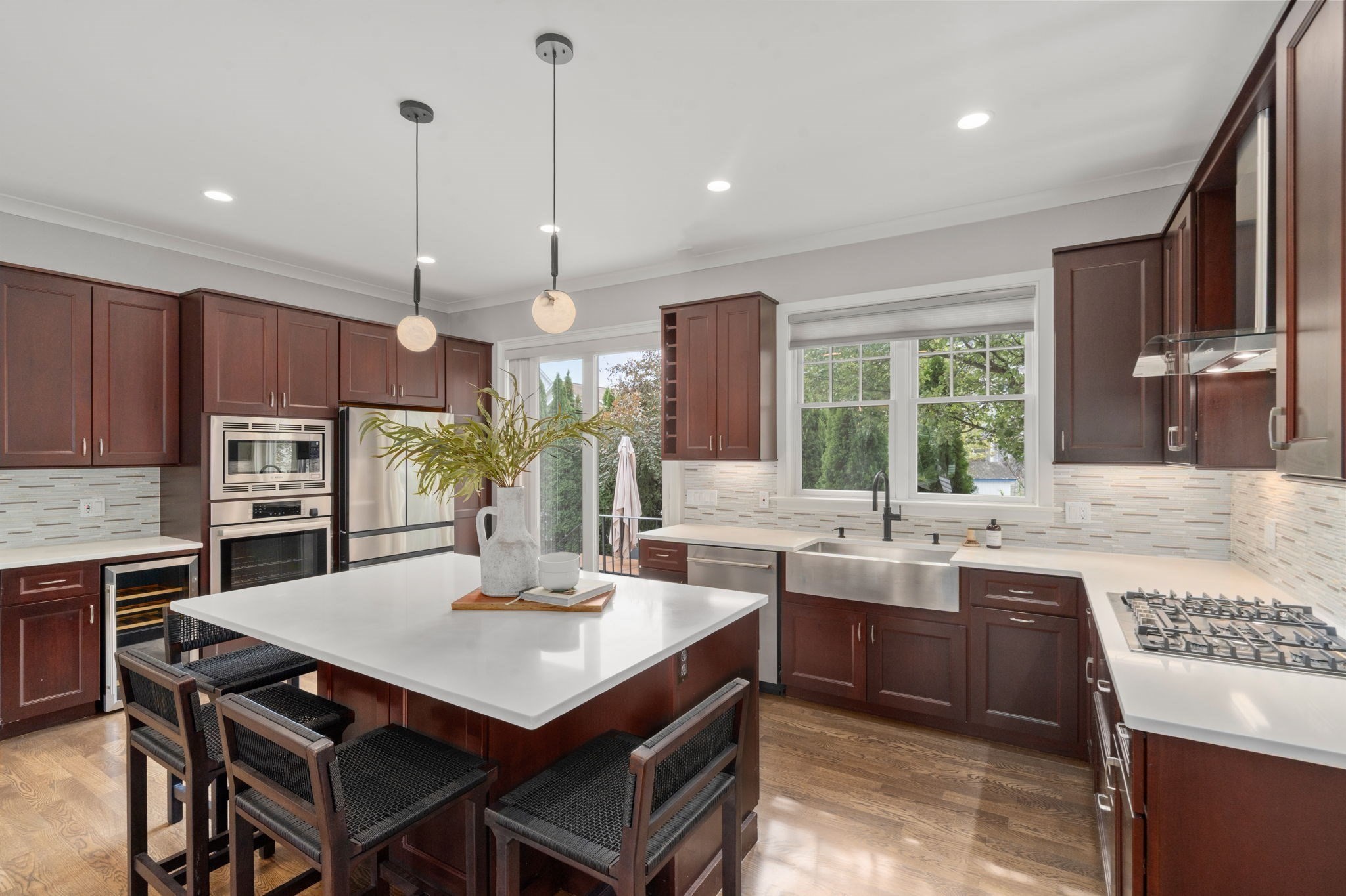 Kitchen with dark cherry cabinets, a large white island, stainless appliances, and a farmhouse sink beneath wide windows.