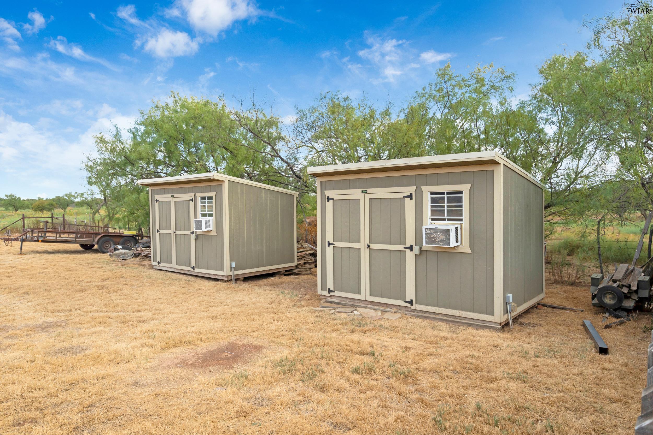 Two beige storage sheds with window air conditioners in a dry, tree-lined yard.