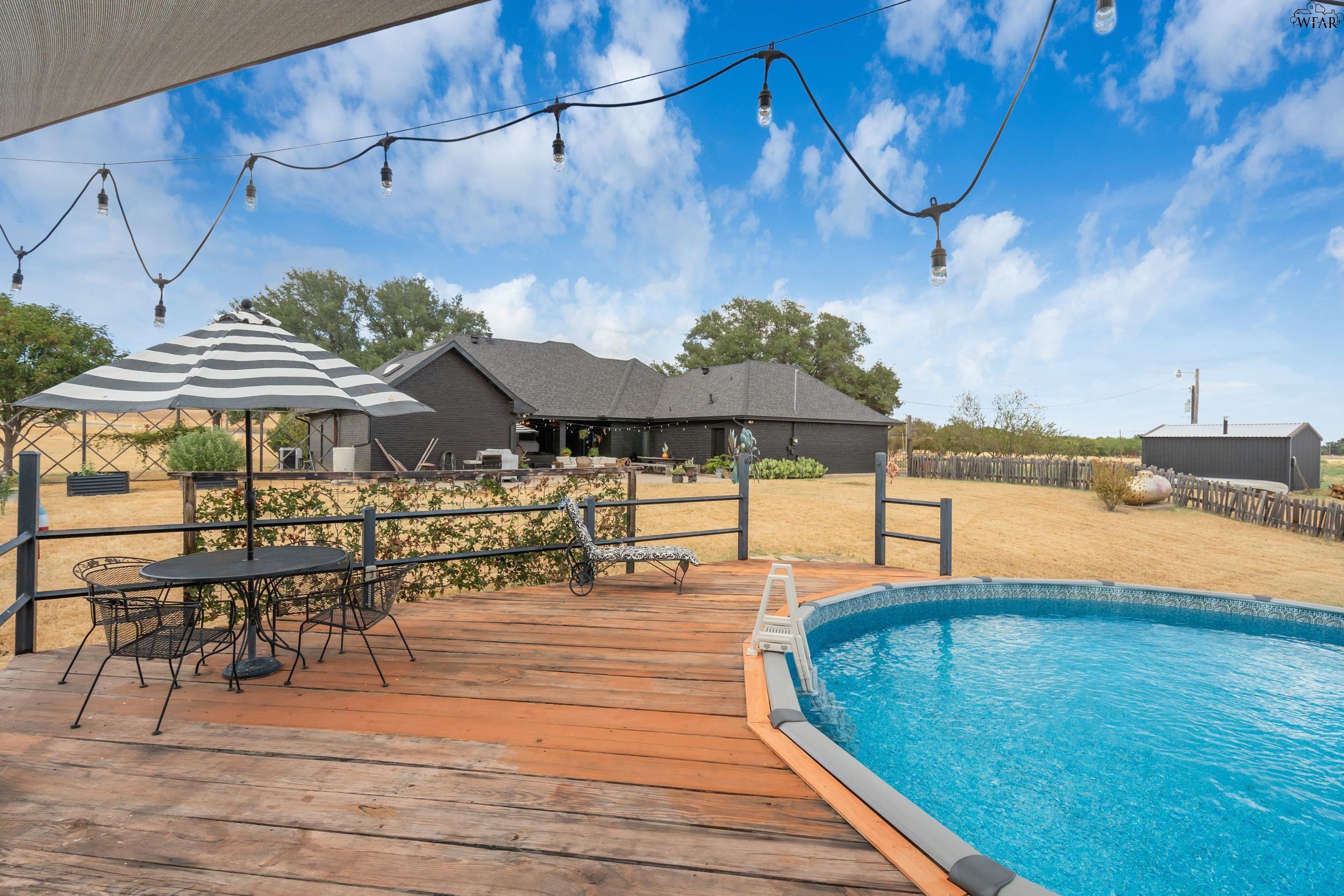 Backyard deck with an above-ground pool, striped umbrella, patio furniture, and string lights.