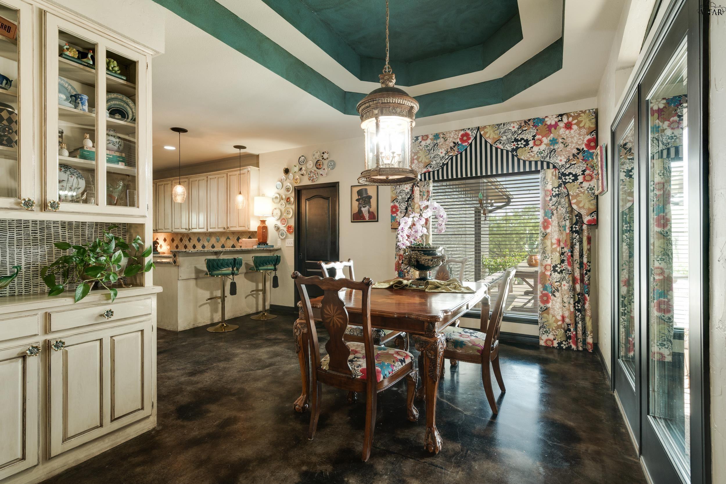 Dining room with a carved wood table, floral chairs, teal tray ceiling, and adjoining kitchen.
