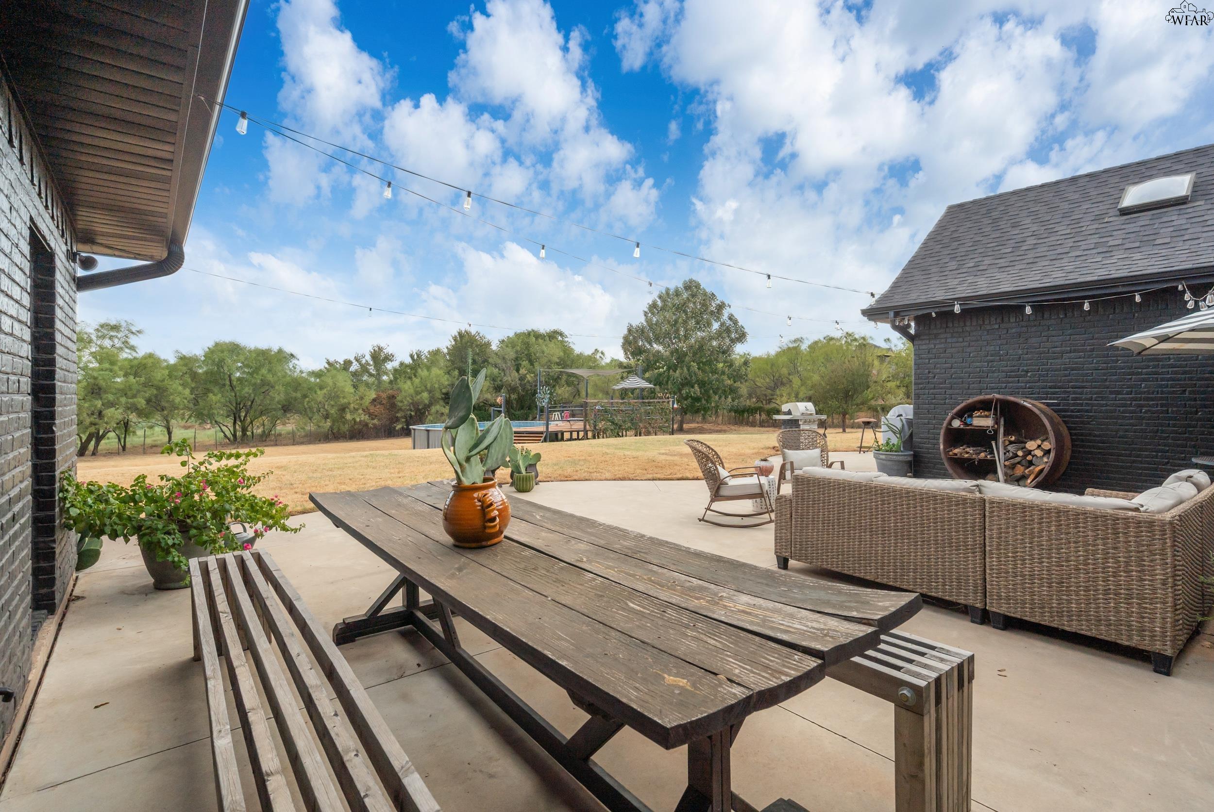 Outdoor patio with a wooden picnic table, woven lounge seating, and string lights overlooking a tree-lined yard.