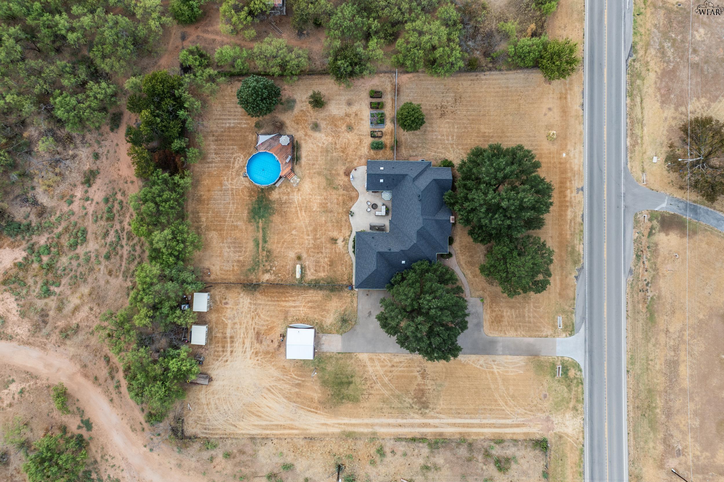 Aerial view of a rural home with a round pool, dry yard, mature trees, and roadside driveway.