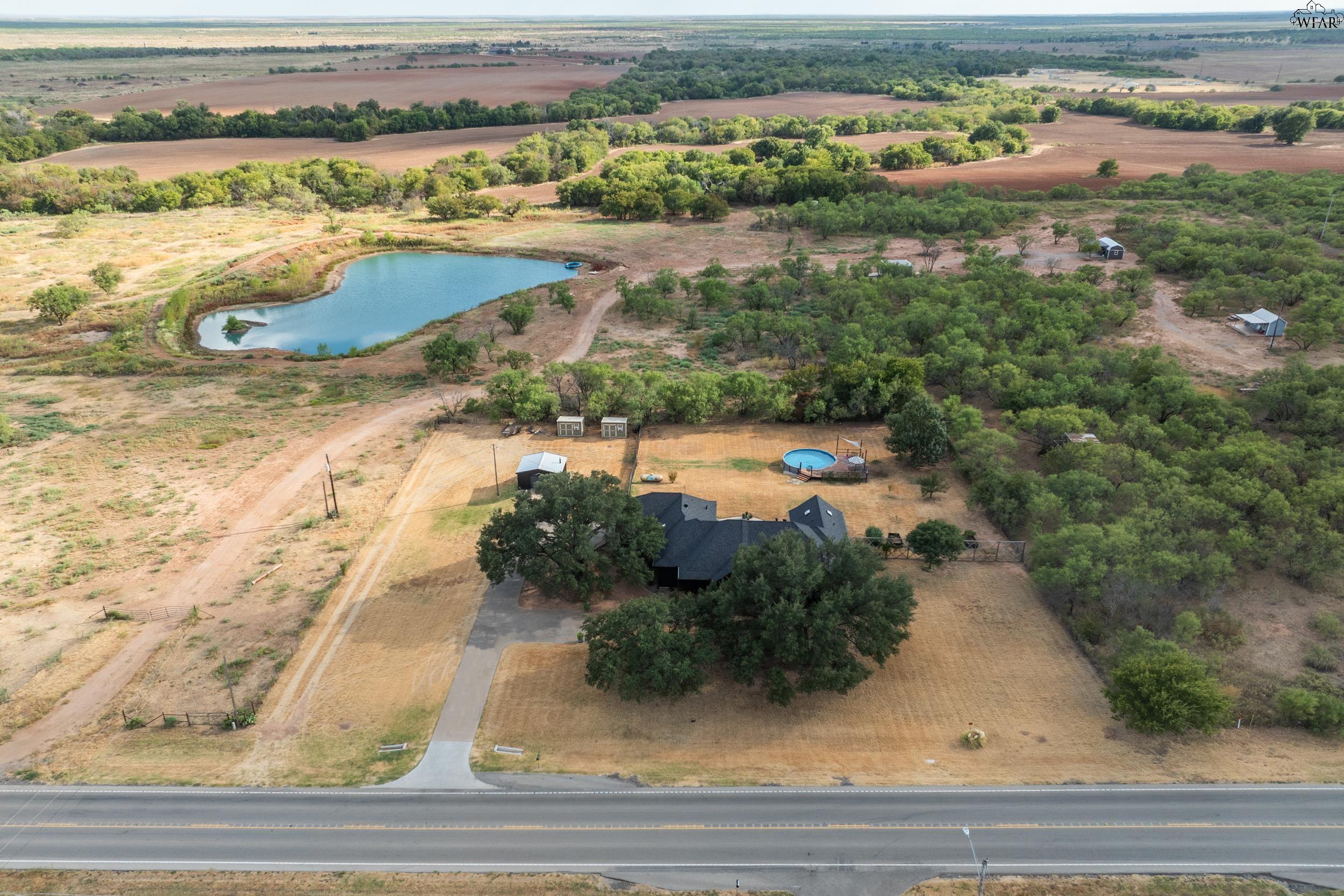 Aerial view of a rural home with a pond, pool, outbuildings, and surrounding fields.