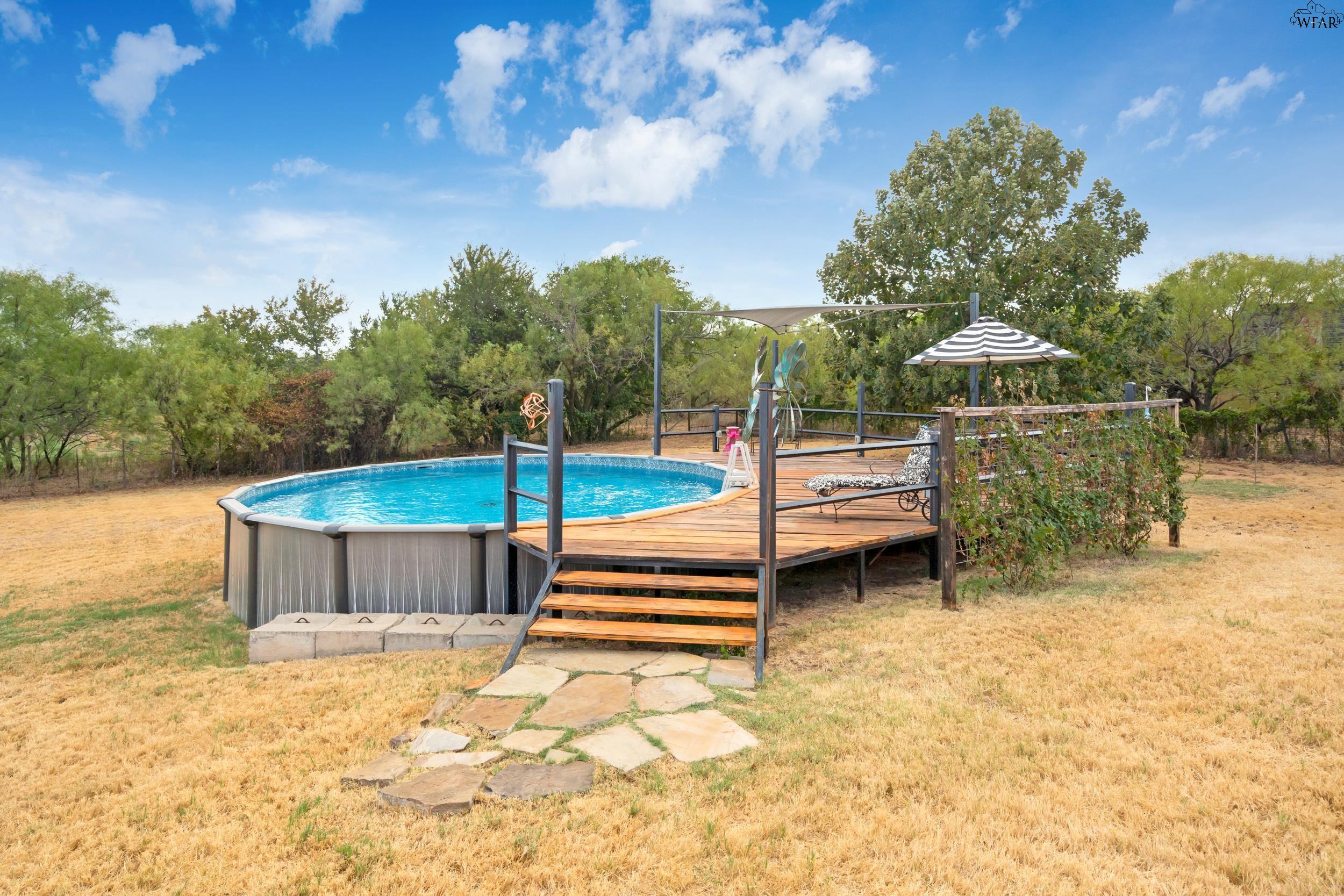 Above-ground pool beside a raised wooden deck with a shade sail and striped umbrella.