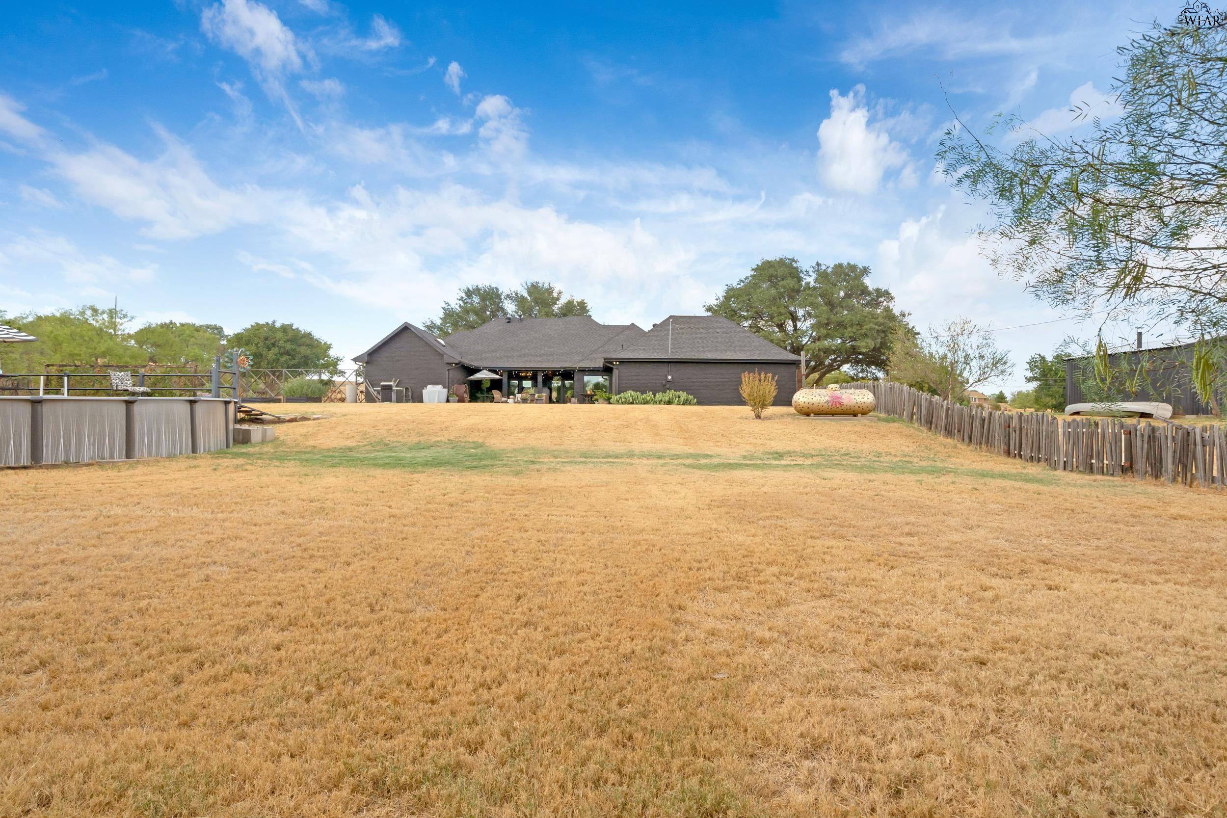 Dark brick home beyond a broad dry lawn, with a raised pool and fenced backyard.