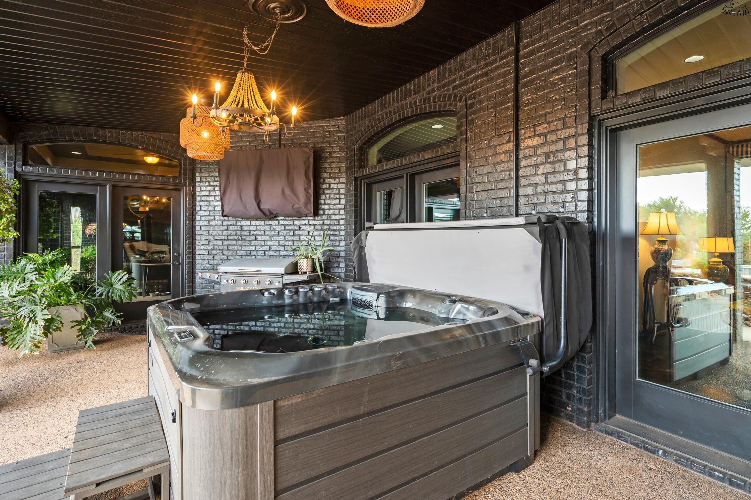 Covered patio with a large hot tub, built-in grill, black brick walls, and chandelier.