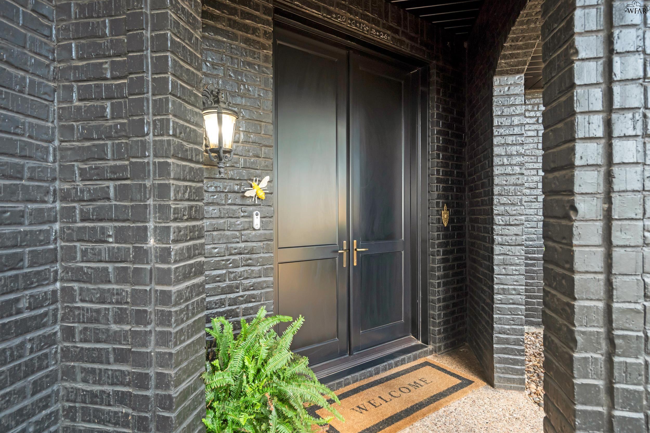 Dark double front doors in charcoal brick, with a glowing lantern, fern, and welcome mat.