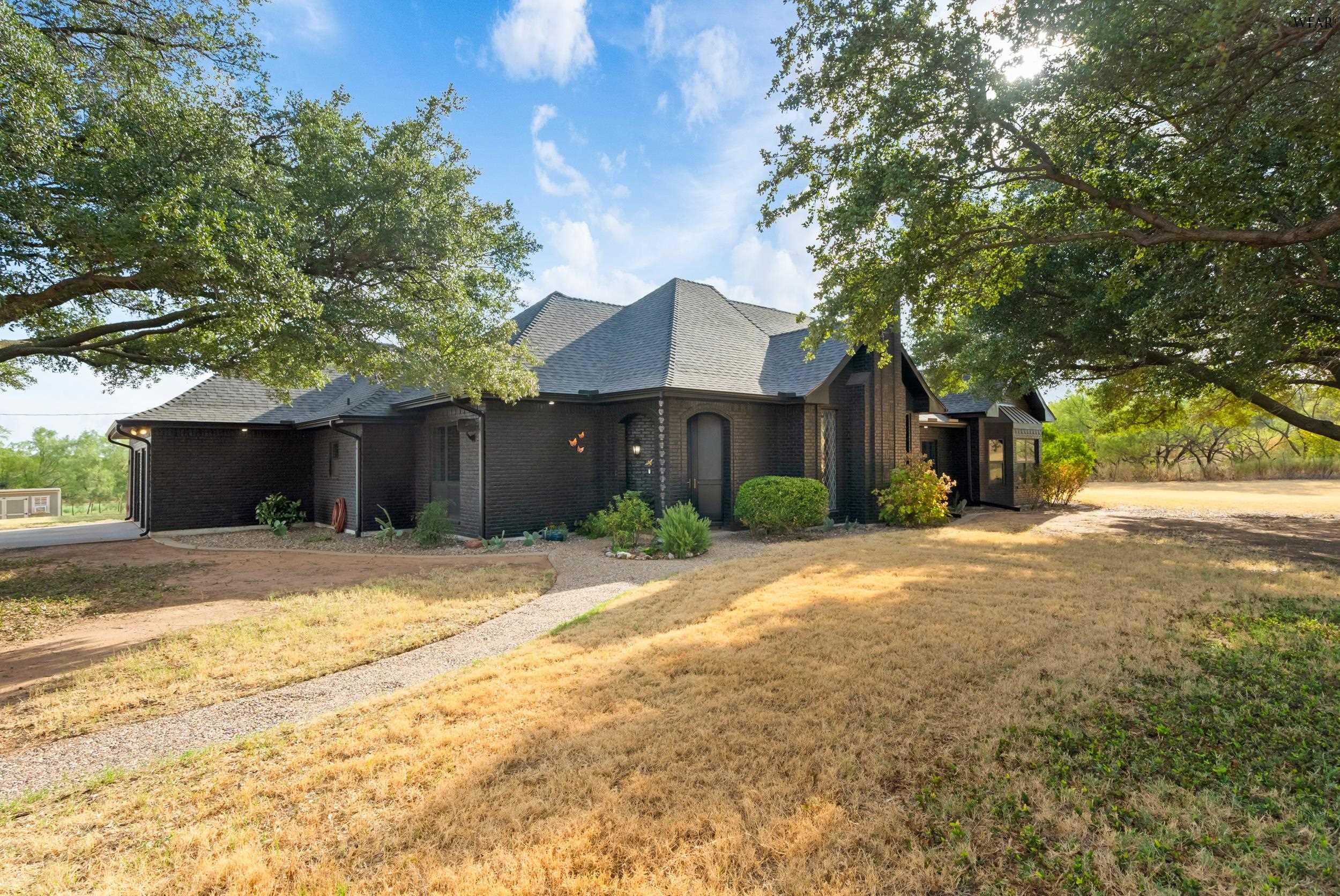 Dark brick country home beneath mature trees, with a gravel path across the dry lawn.