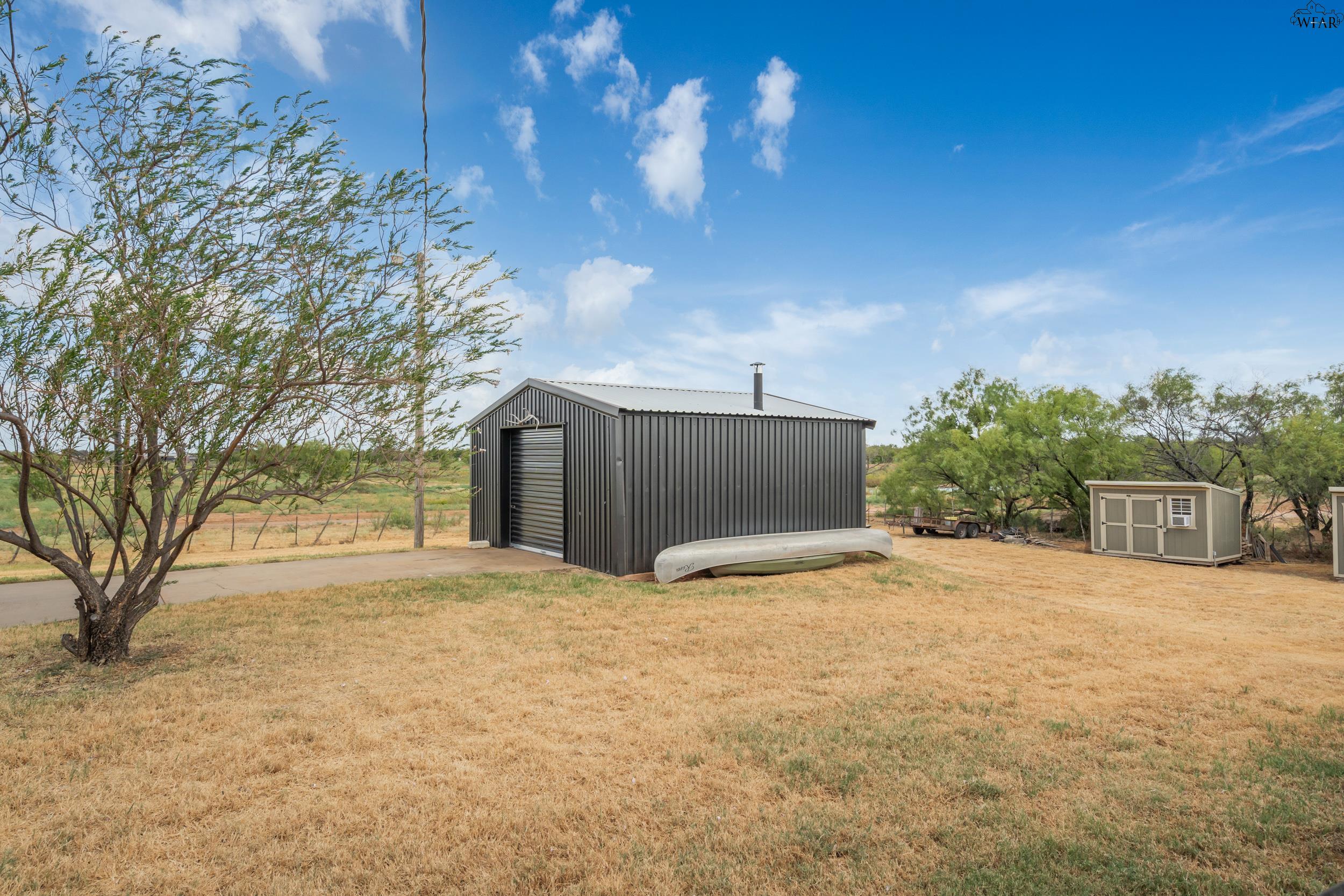 Rural property with a dark metal outbuilding, small shed, and overturned canoe on dry grass.
