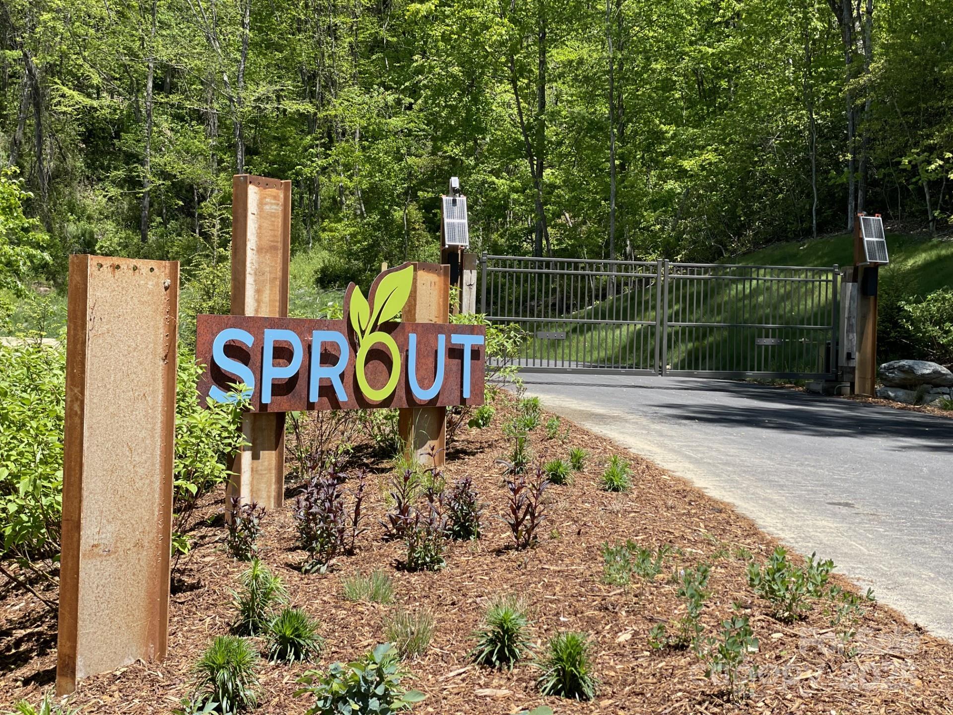 Sprout community entrance sign beside a metal gate, landscaped beds, and wooded hillside.