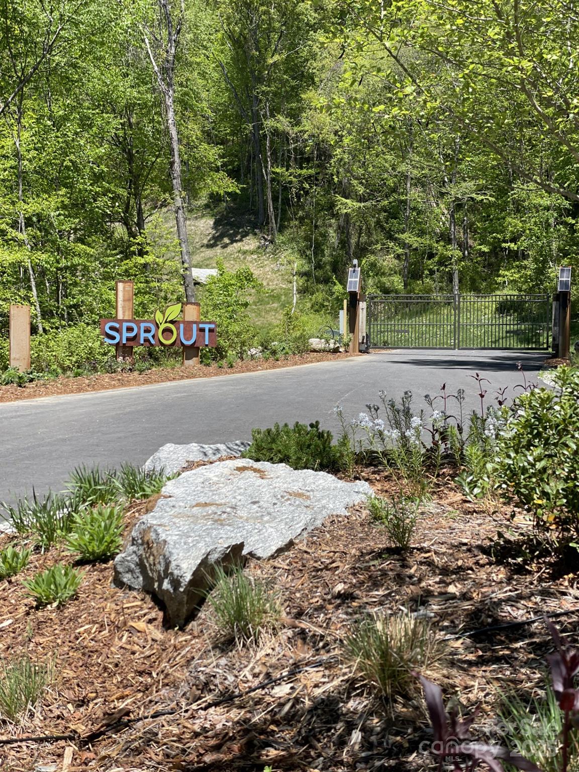 Sprout community entrance with a metal gate, wooded hillside, and landscaped roadside.