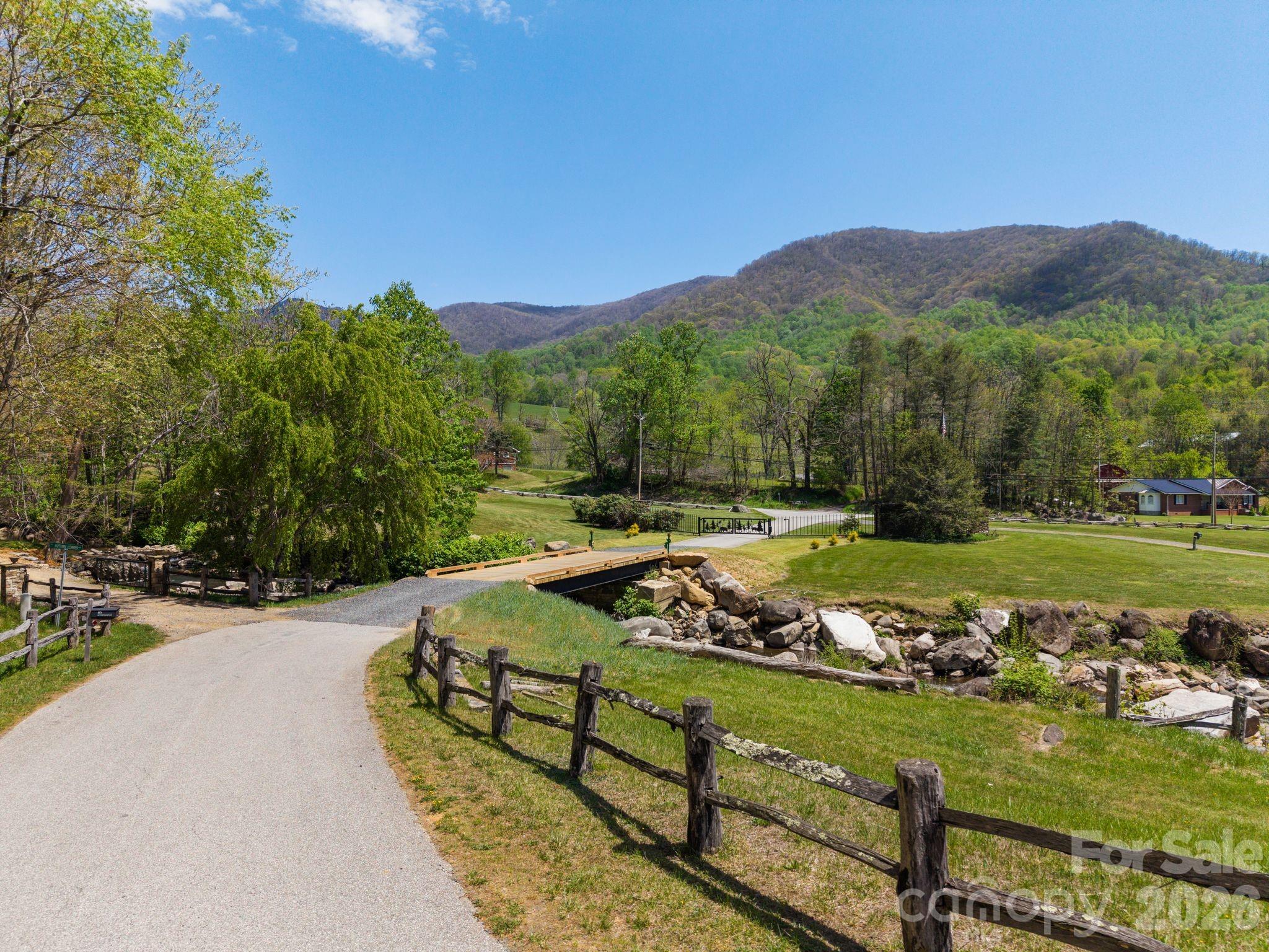 Landscaped community lane and creek bridge with wooded mountains beyond.