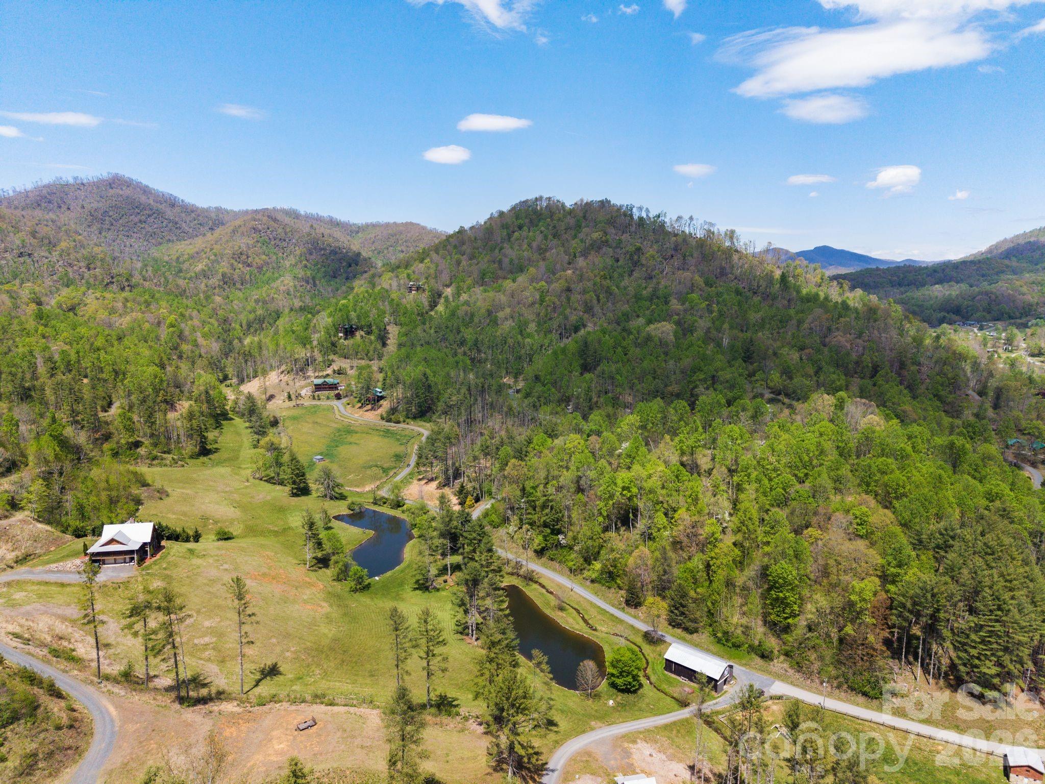 Aerial mountain property with wooded hills, open fields, scattered buildings, winding roads, and two ponds.
