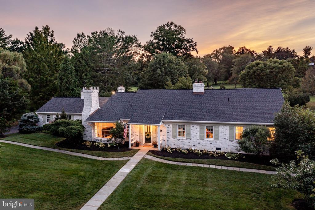 White brick home with a dark roof and glowing windows, framed by a landscaped lawn at dusk.