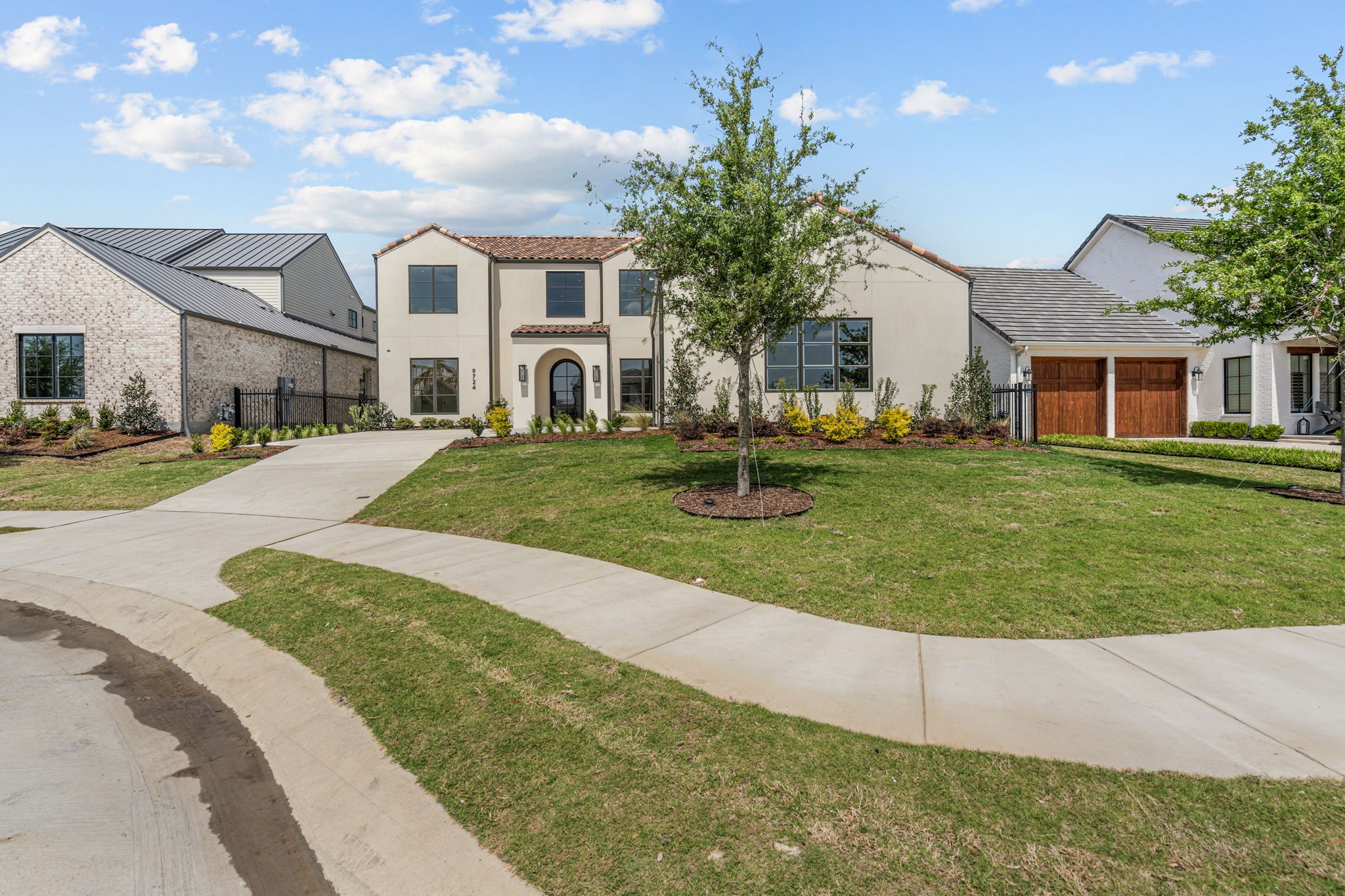 Cream-colored home with an arched entry, tile roof, wooden garage doors, and landscaped front lawn.