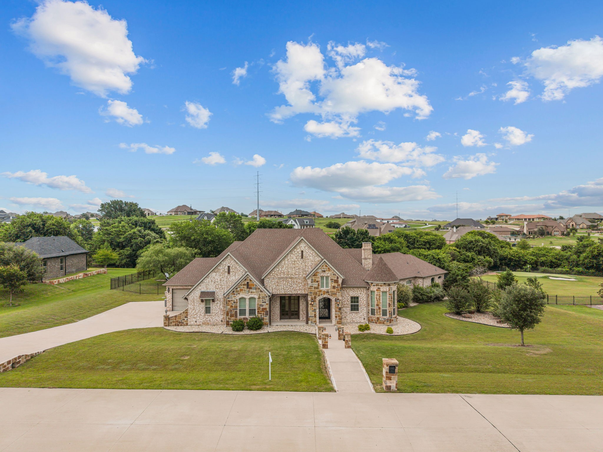Stone-and-brick house with a broad lawn, curved driveway, and neighboring homes in the distance.