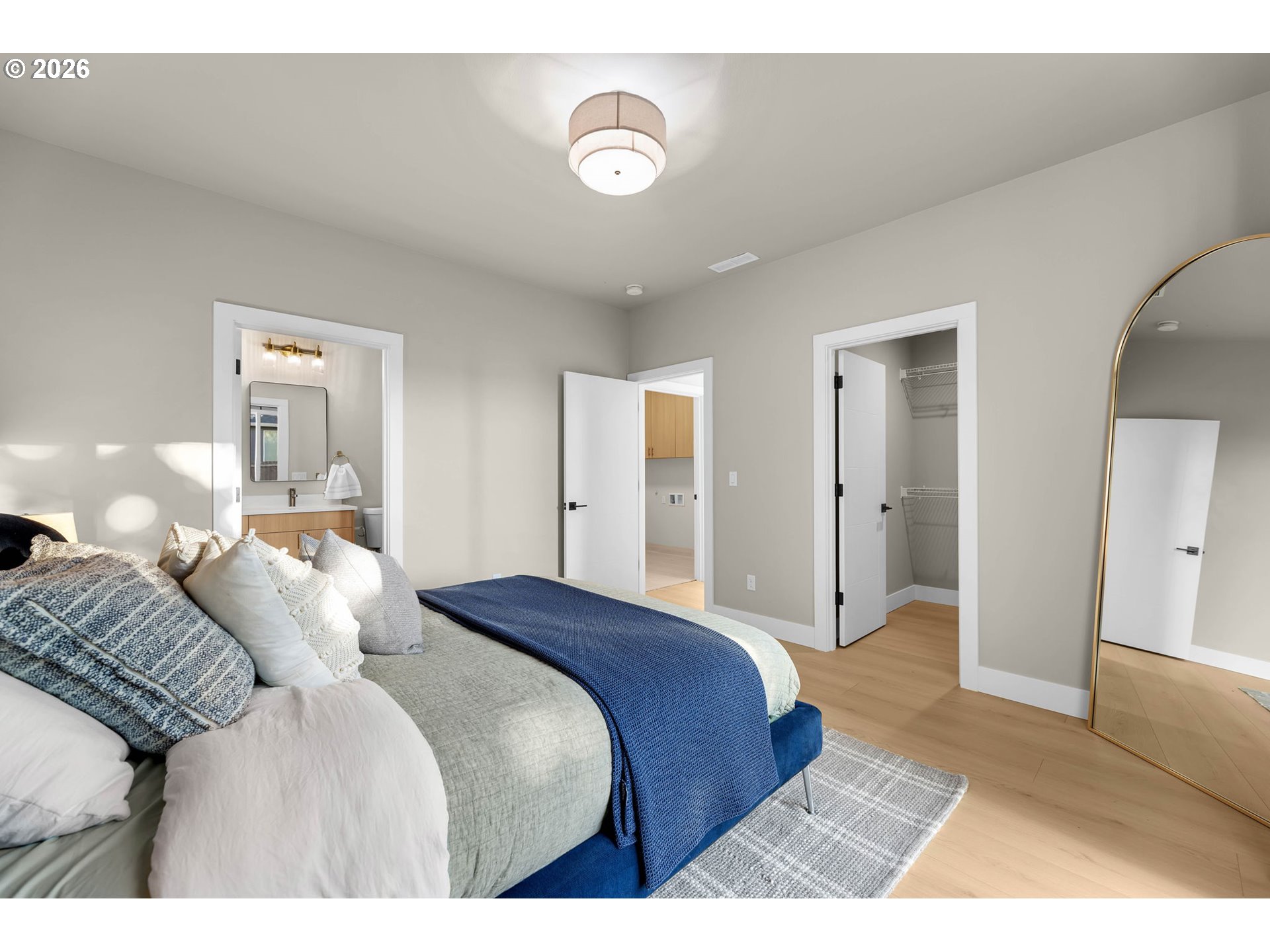 Staged bedroom with blue bedding, an arched floor mirror, and open ensuite and closet doors.