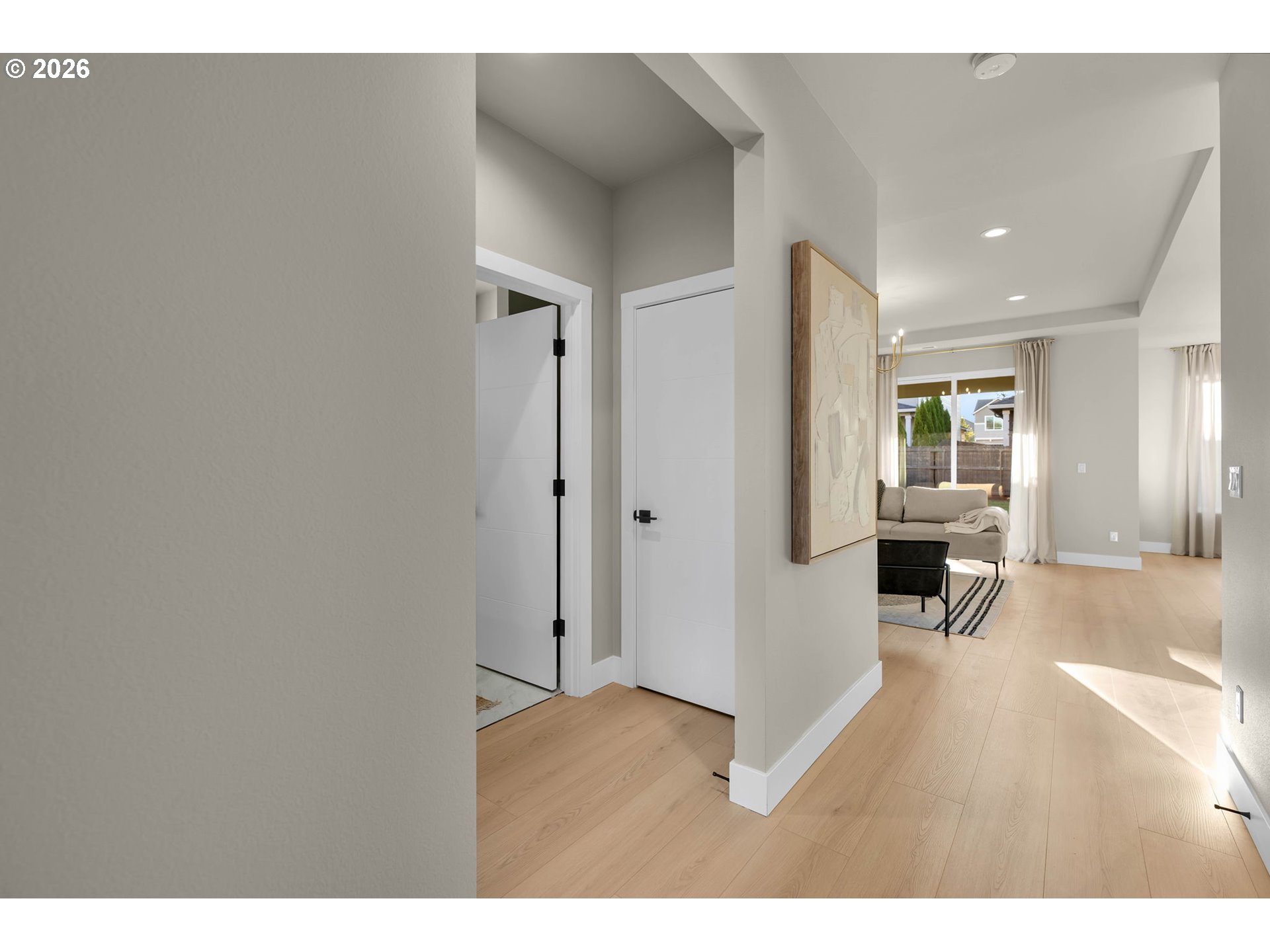 Hallway with light wood floors opening into a living room with a neutral sofa and large windows.