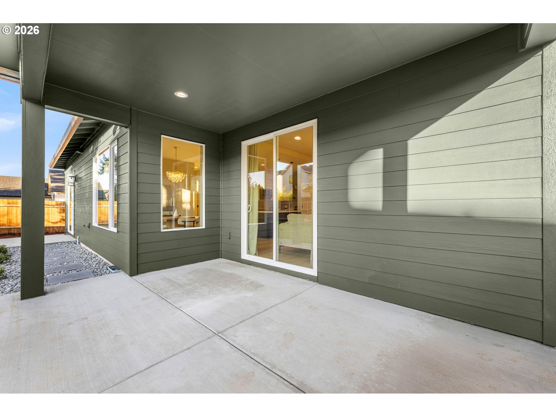 Covered concrete patio beside a modern gray-sided home, with a sliding glass door and gravel walkway.