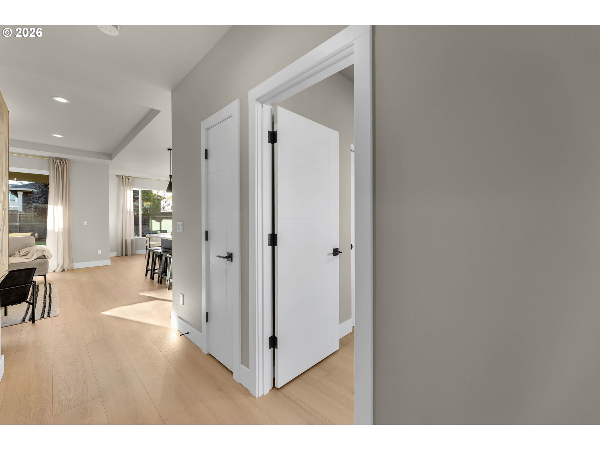 Hallway with light wood floors and white doors opening toward a sunlit living and dining area.
