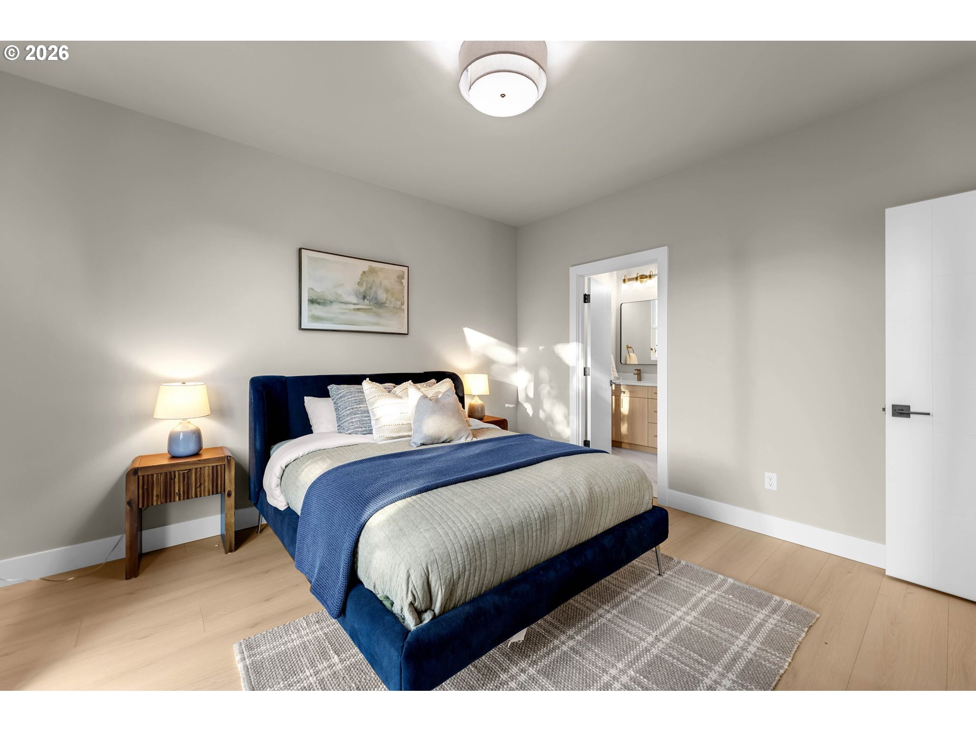 Staged bedroom with a navy bed, blue throw, light wood floors, and an adjoining bathroom doorway.