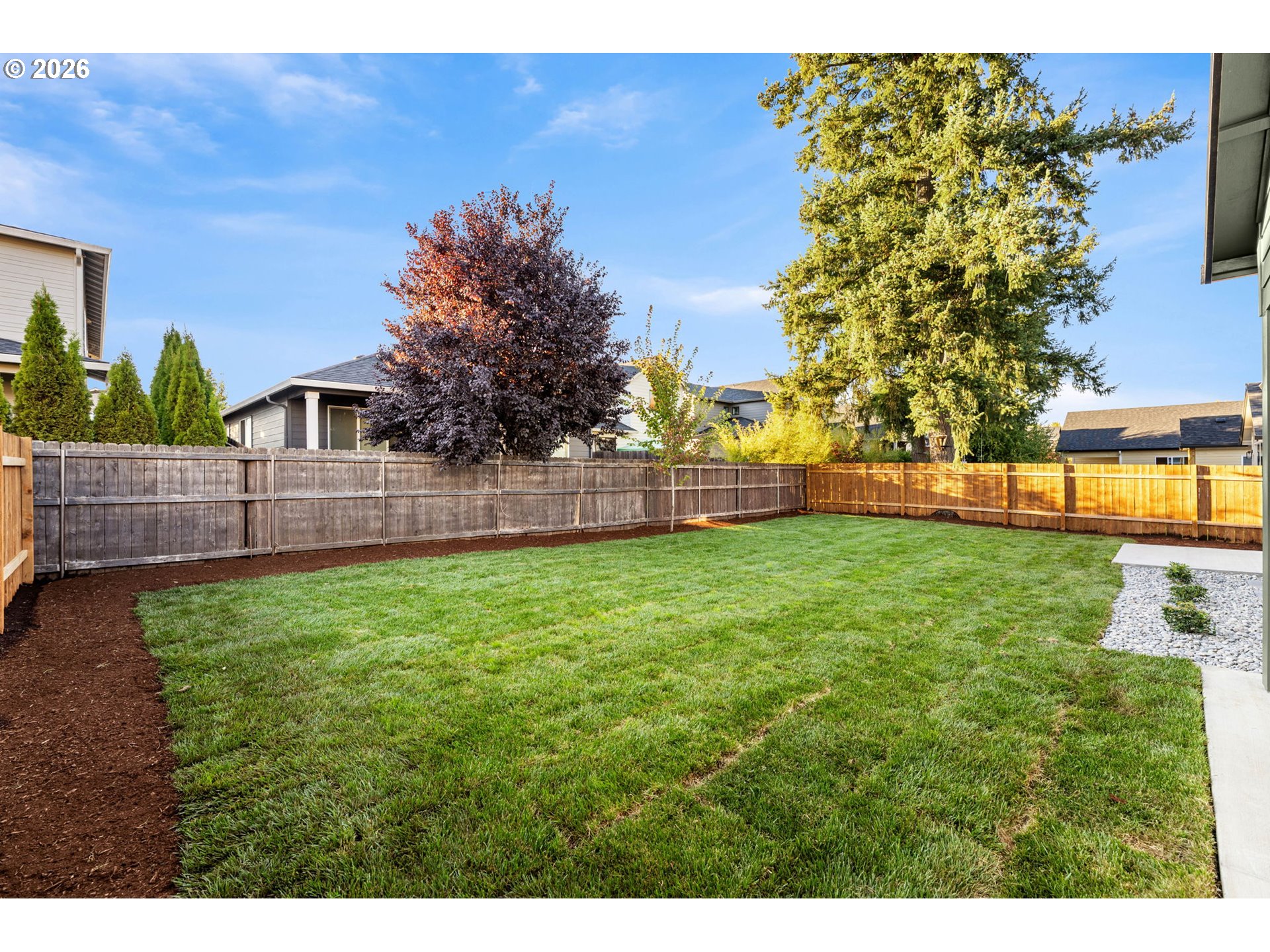 Fenced backyard with a broad lawn, mature trees, and a white-stone landscaping area.