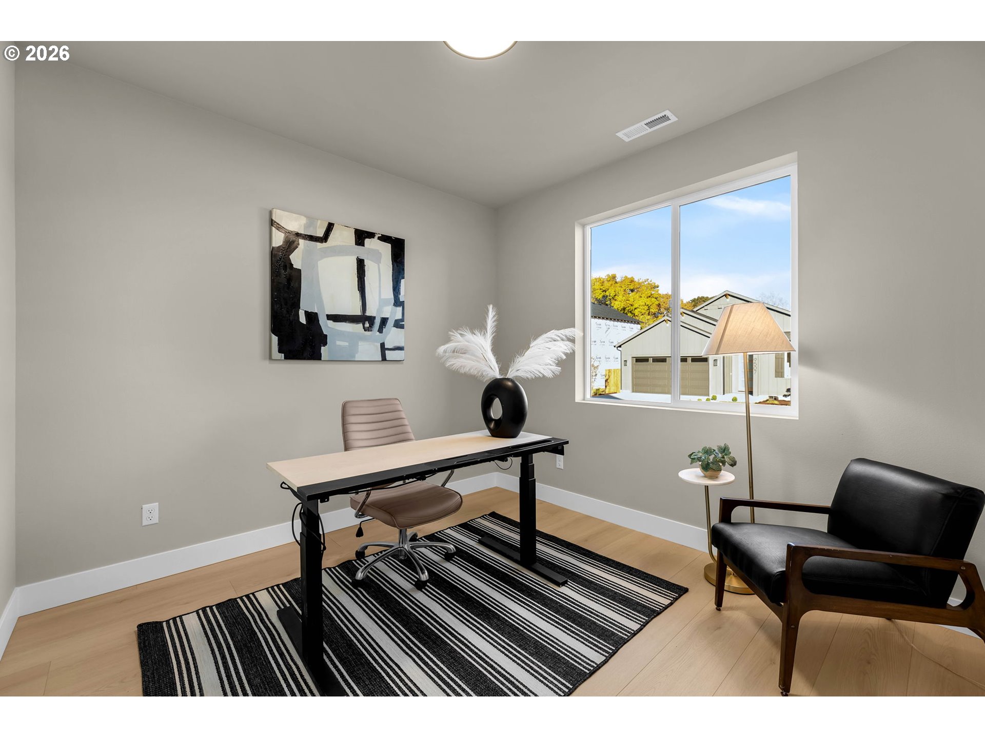 Furnished home office with a wood desk, striped rug, black armchair, and large window.