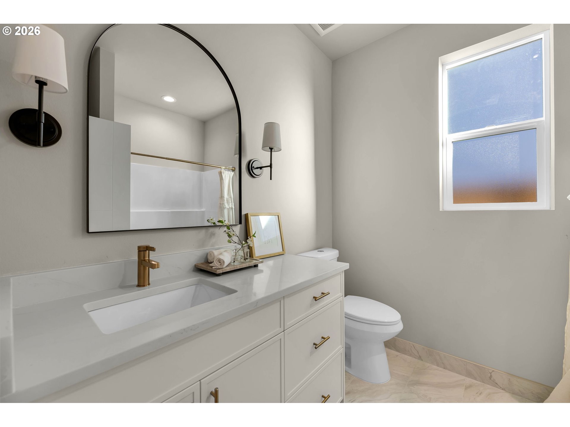 Bathroom with a white vanity, brass fixtures, arched mirror, and window.