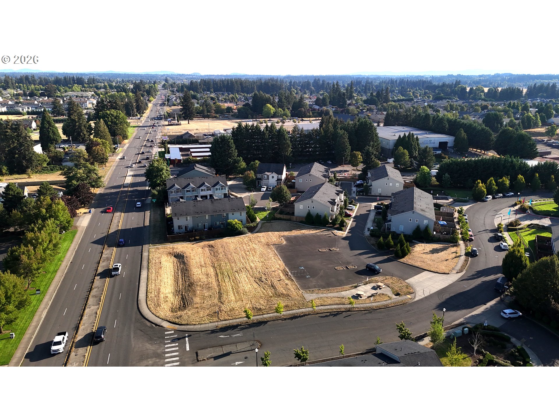 Aerial view of a vacant corner lot beside a main road, with nearby homes and mature trees.