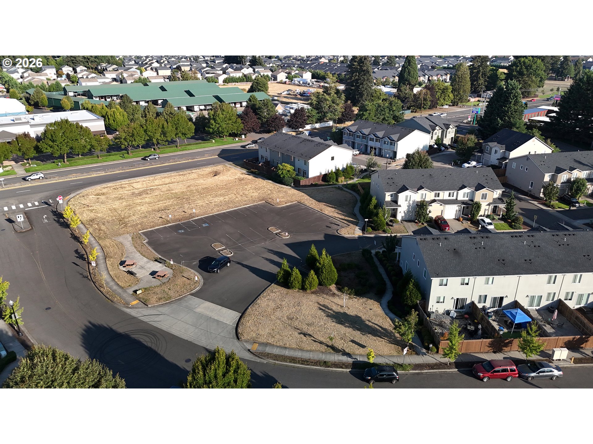 Aerial view of a vacant lot beside a road, with a paved parking area and nearby townhomes.