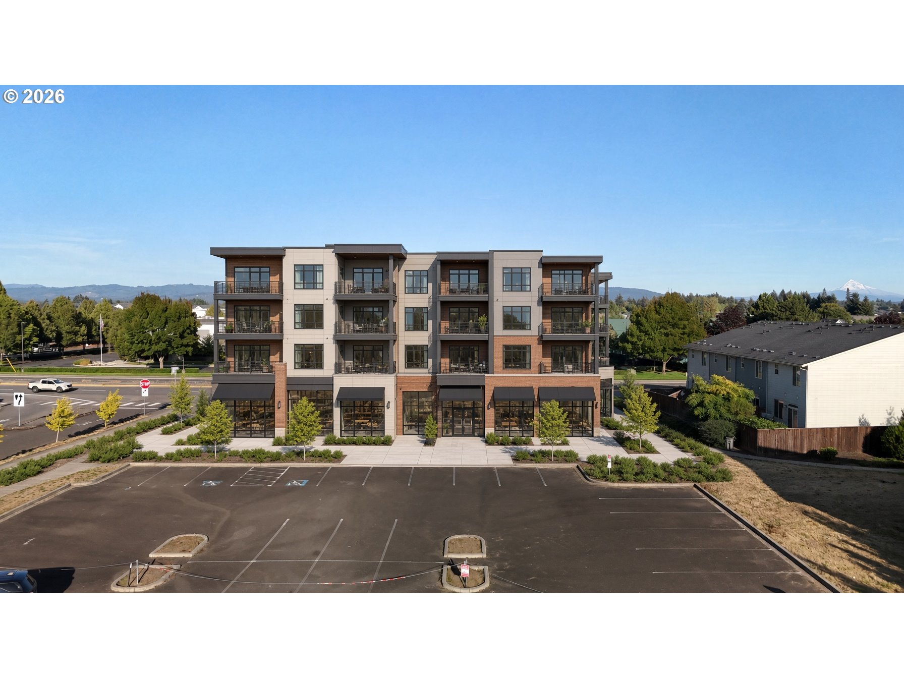 Modern multi-story building with balconies, a broad parking lot, and a distant snow-capped mountain.