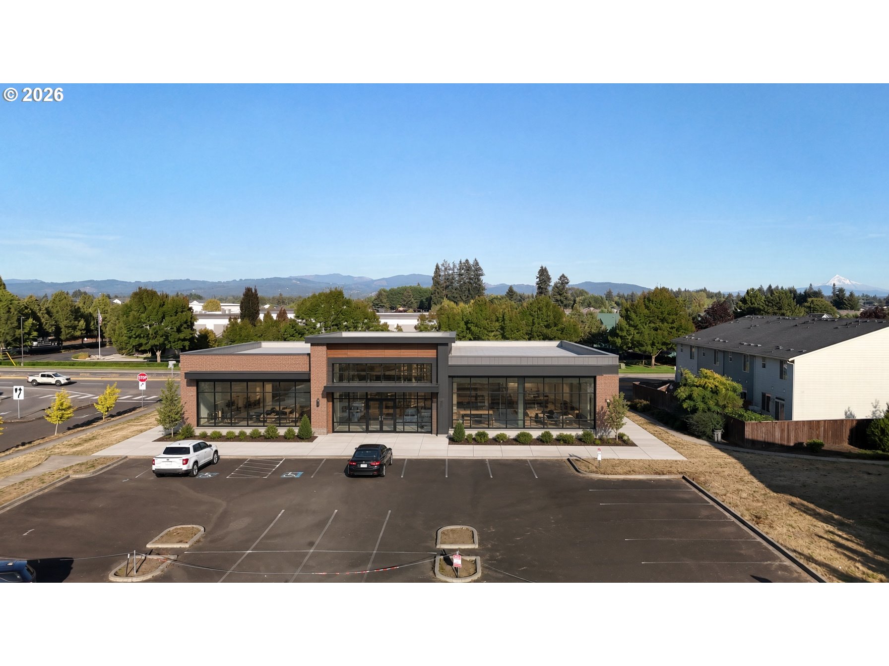 Modern commercial building with brick, broad glass windows, and a parking lot, backed by distant hills and a snow-capped peak.