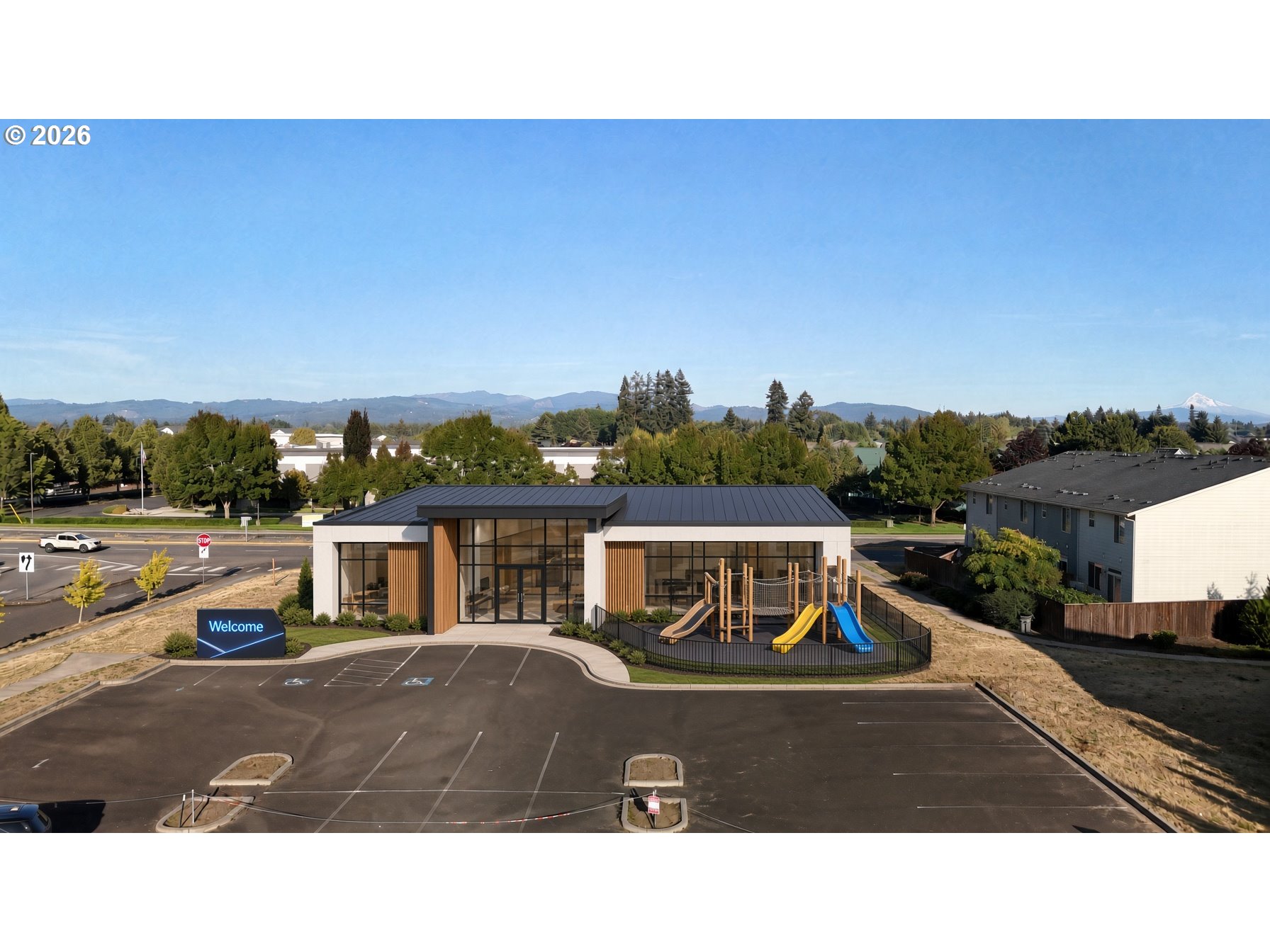 Modern building with a fenced playground and parking lot, backed by distant mountains.
