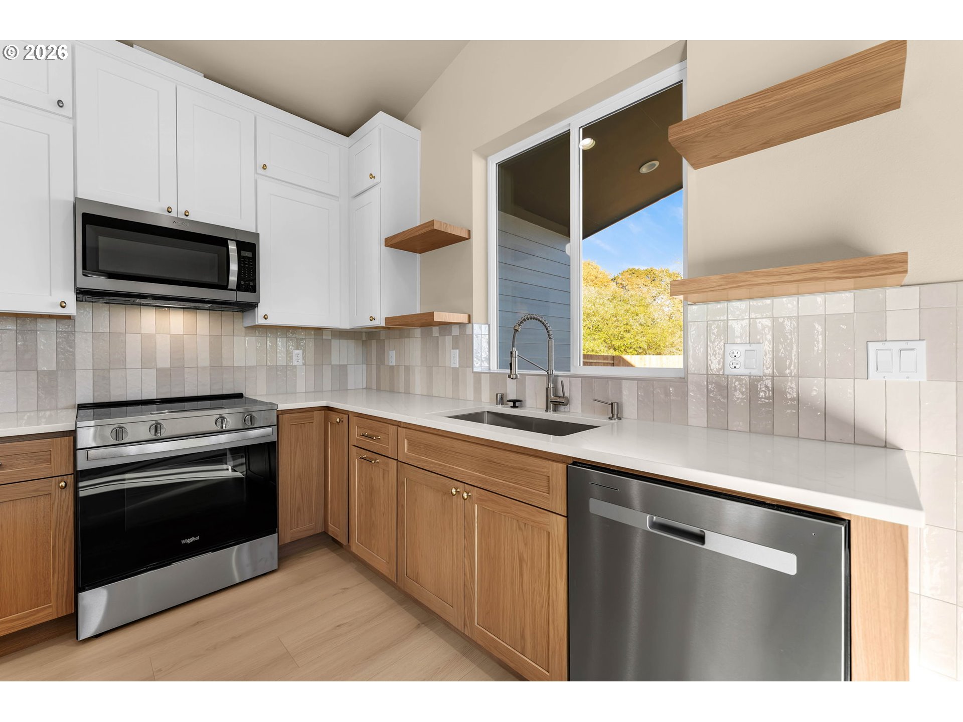 Modern kitchen with wood cabinets, floating shelves, stainless steel appliances, and a large window above the sink.