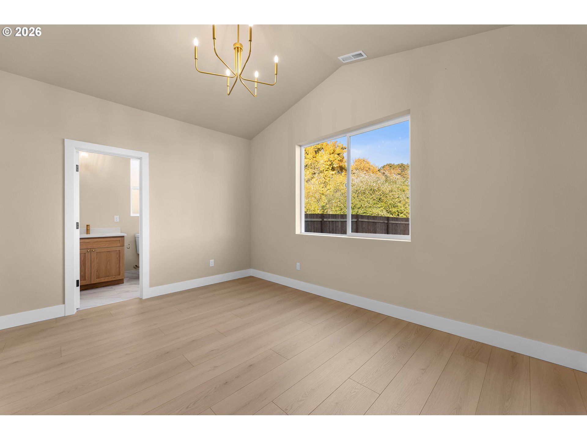 Empty bedroom with light wood flooring, a brass chandelier, and a window overlooking autumn trees.