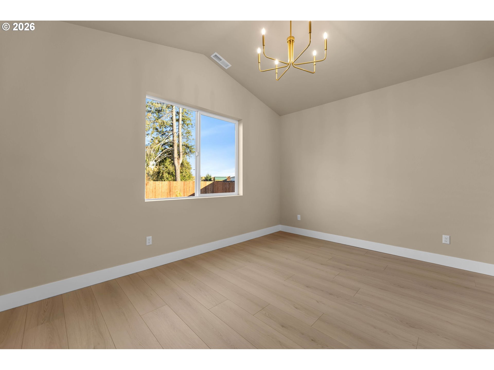 Vacant room with vaulted ceiling, light wood floors, large window, and gold chandelier.