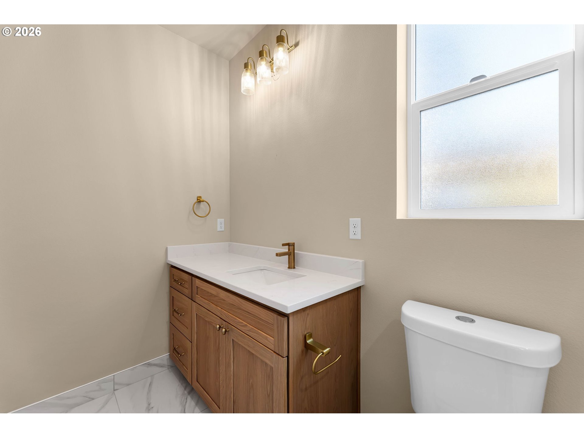 Bathroom vanity with a wood cabinet, brass fixtures, frosted window, and white toilet.