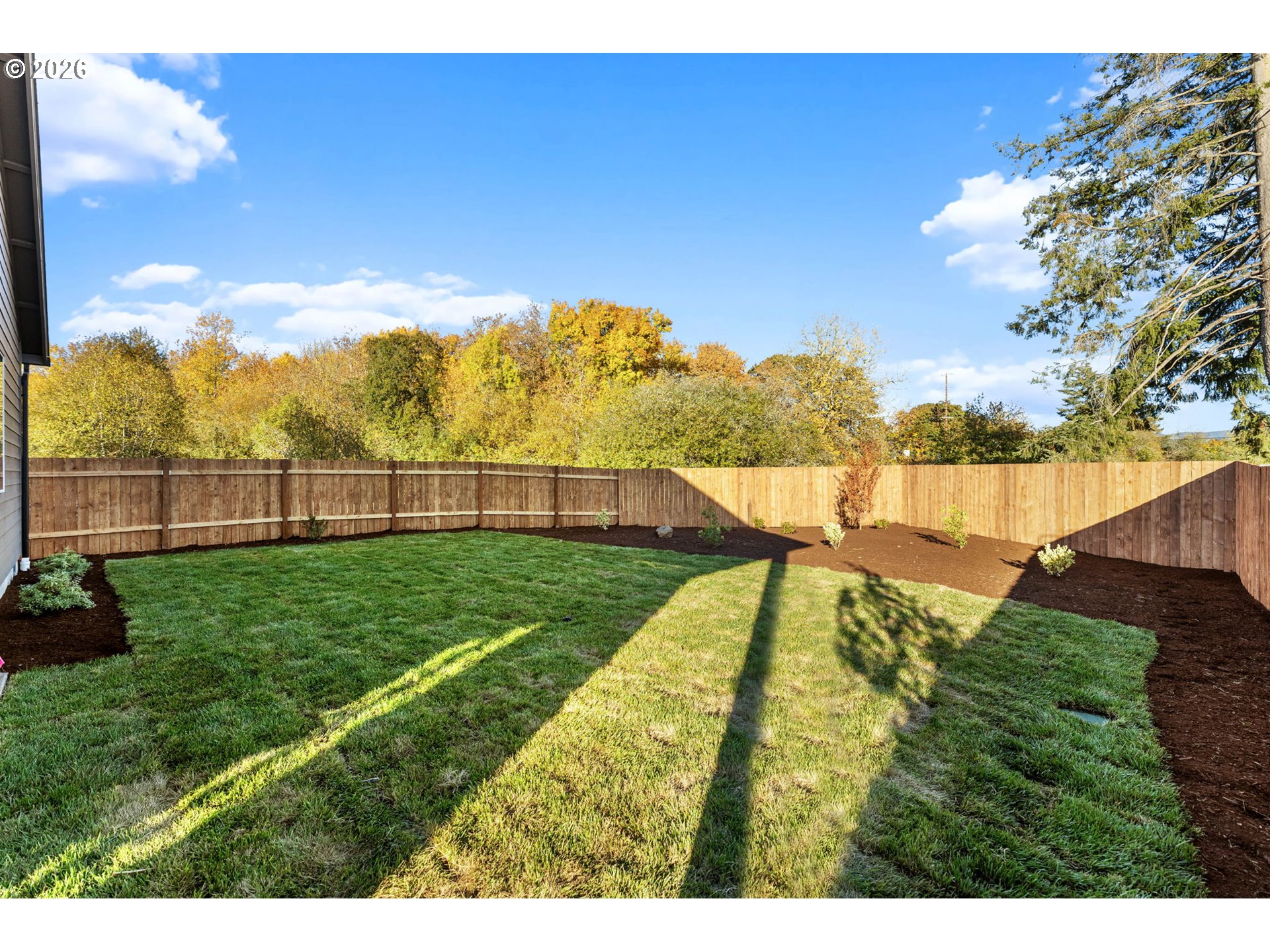 Fenced backyard with a green lawn, mulched beds, and autumn trees beyond the fence.