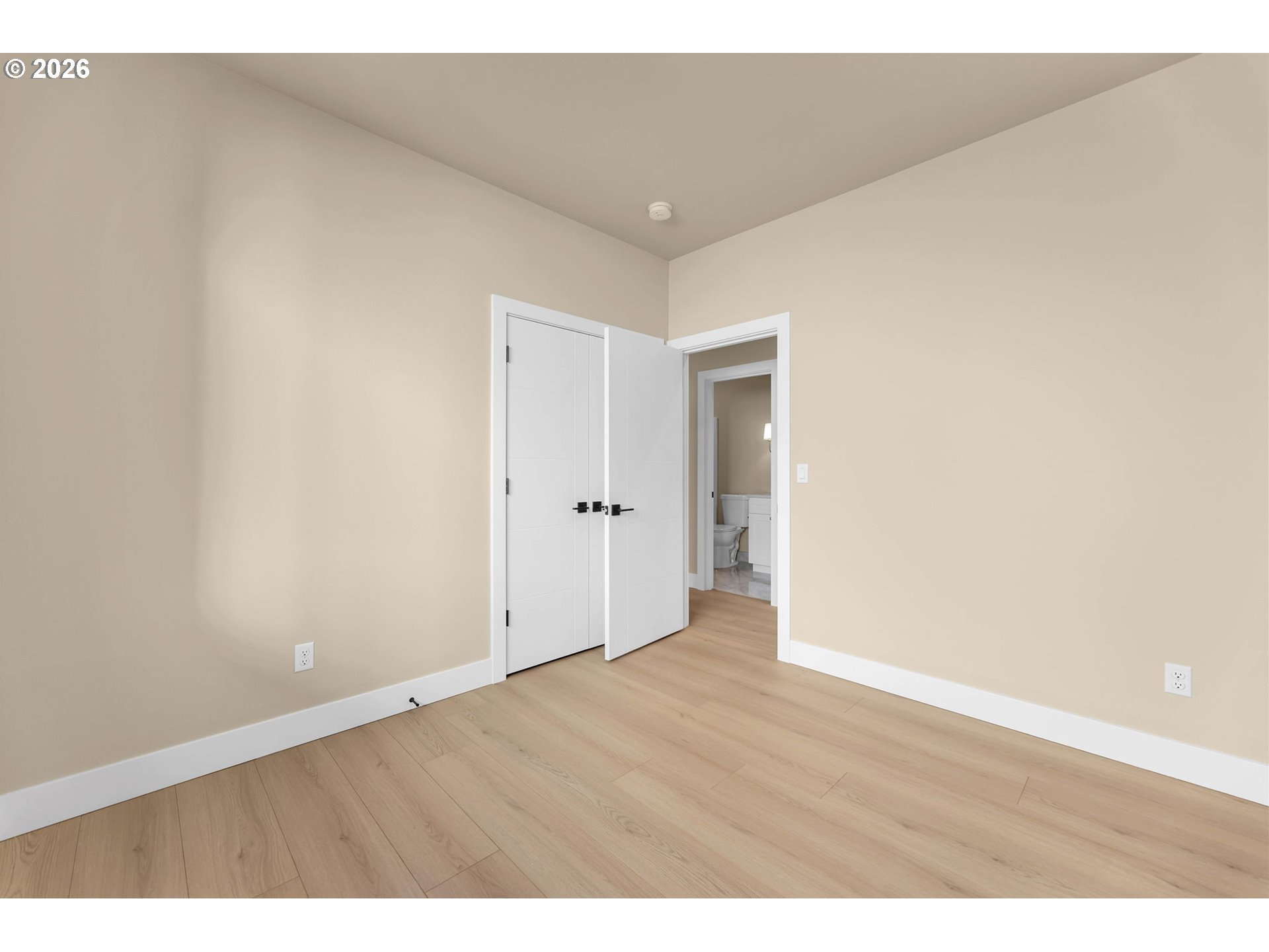 Empty bedroom with light wood floors and open doors to a bathroom.